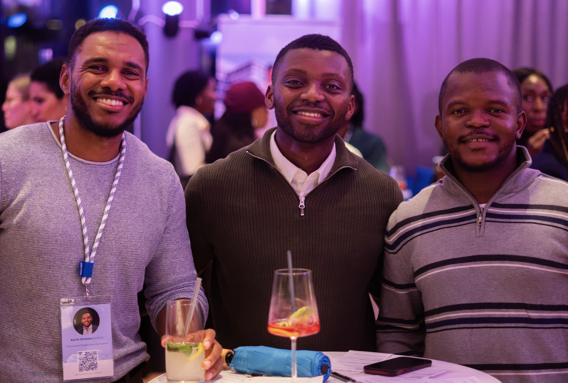 Three smiling men standing together at a social event, with drinks on the table in front of them, purple and pink lighting, blurred background with other attendees.