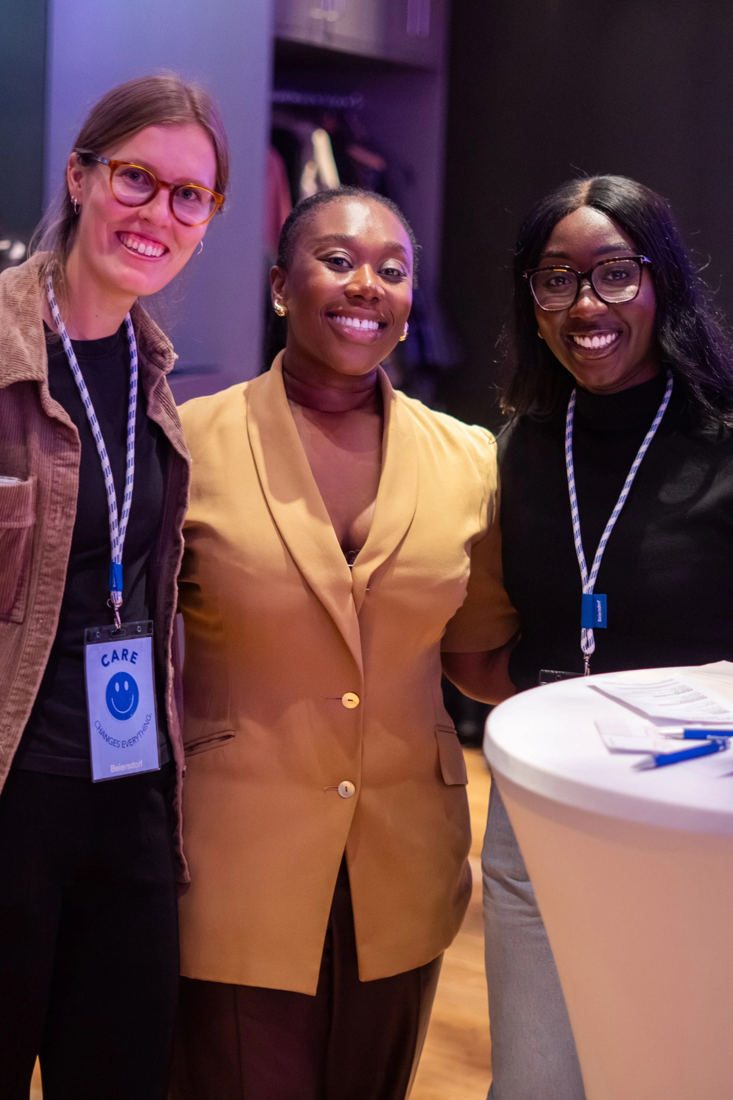 Three women standing together at an indoor event, smiling at the camera. The woman on the left has light skin, glasses, and is wearing a brown jacket. The woman in the middle has dark skin, is wearing a tan blazer, and has earrings. The woman on the right has dark skin, glasses, and is wearing a black top. There is a high table with papers and a pen in the foreground.