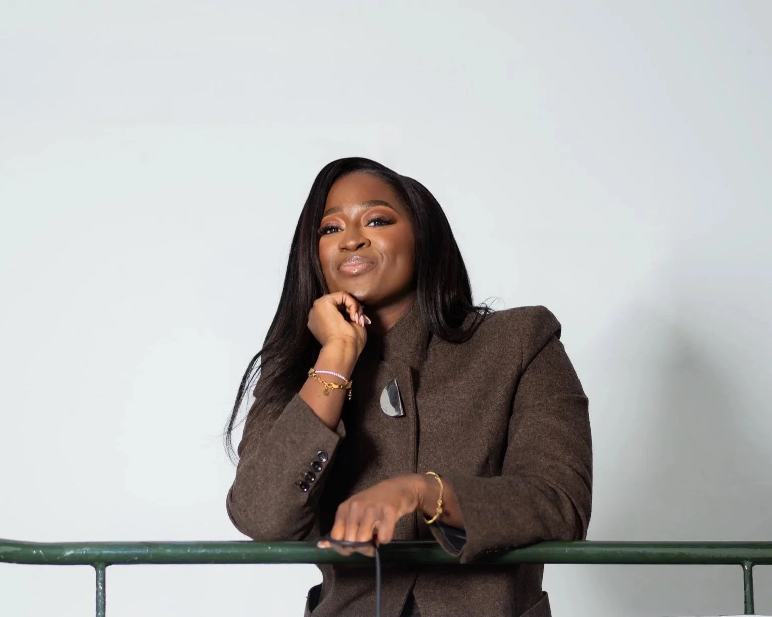 A woman with long black hair wearing a brown blazer smiles while resting her chin on her hand, standing against a plain light grey background.