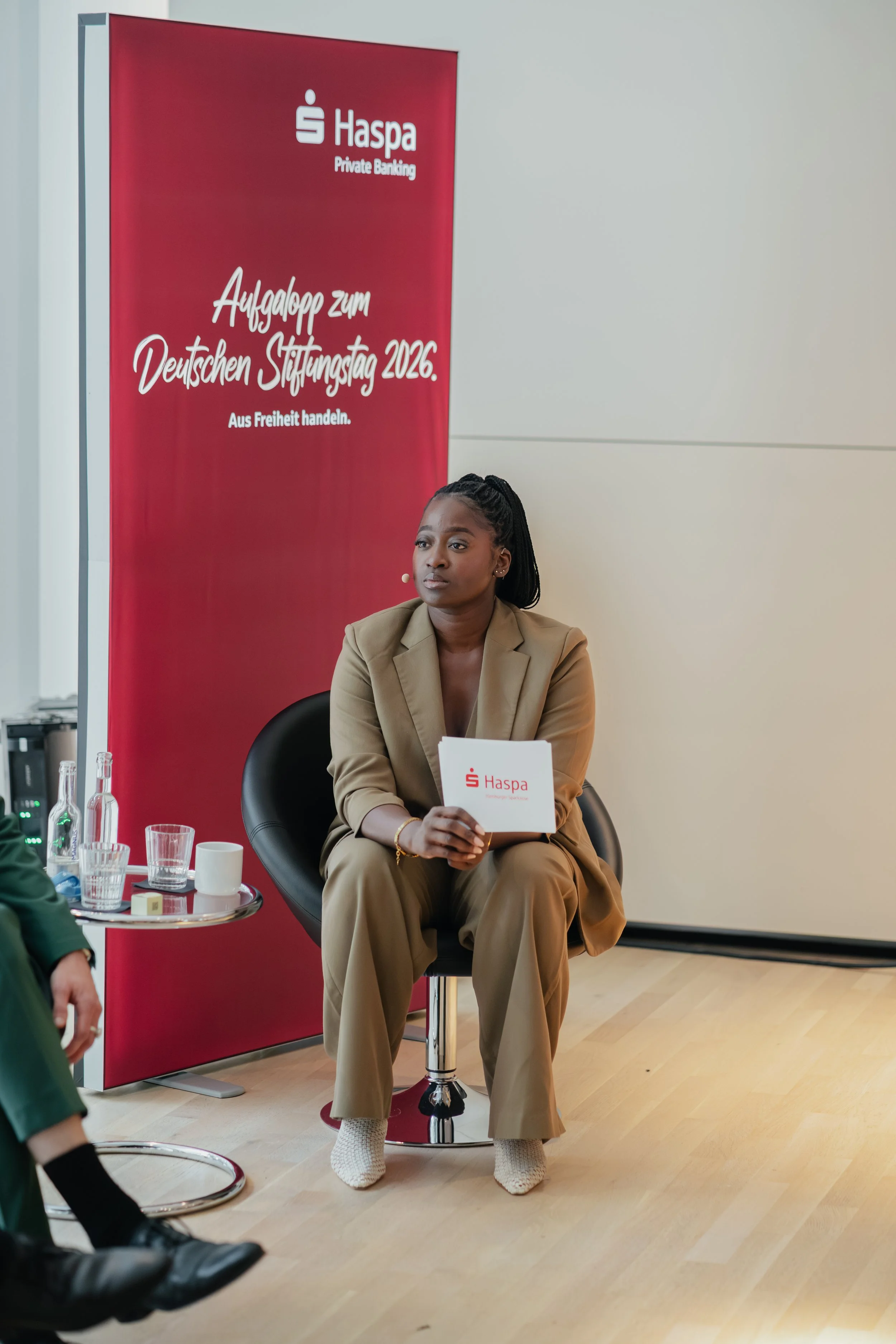 A woman in beige pants and blazer sitting on a chair, holding a Haspa brochure, at a table with beverages, with a red Haspa banner behind her that reads "Aufgalopp zum Deutschen Stiftungstag 2026" and "Aus Freiheit handeln."