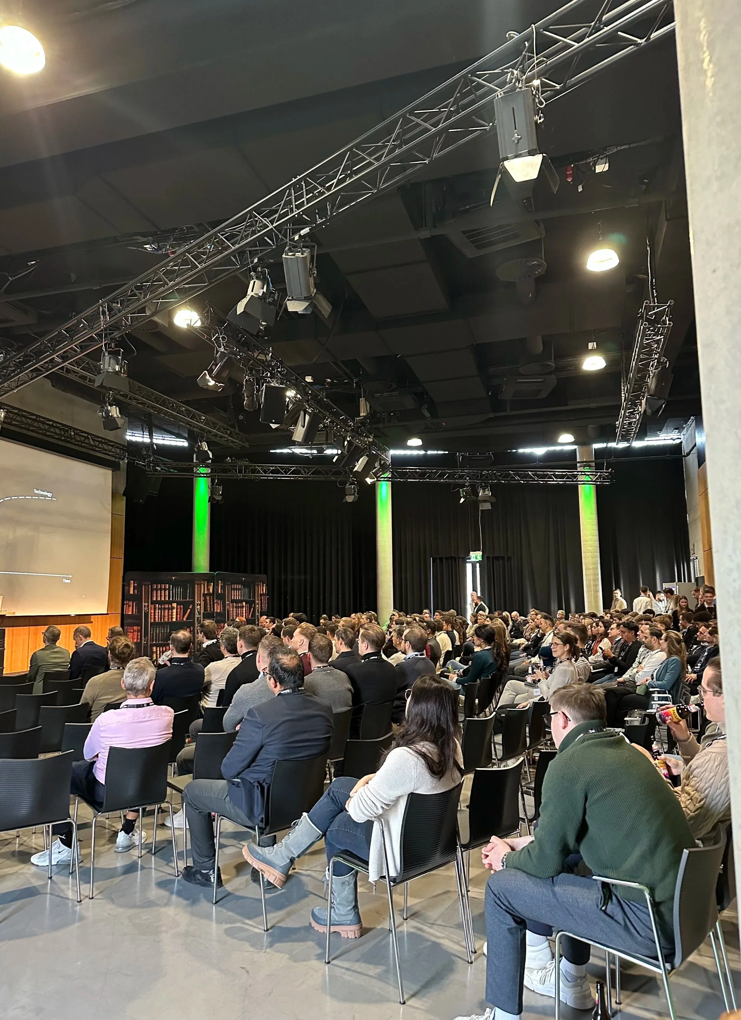 A large audience seated in rows of chairs in a conference room, facing a stage or presentation area, with some individuals taking notes or using phones, and a few standing near the back.
