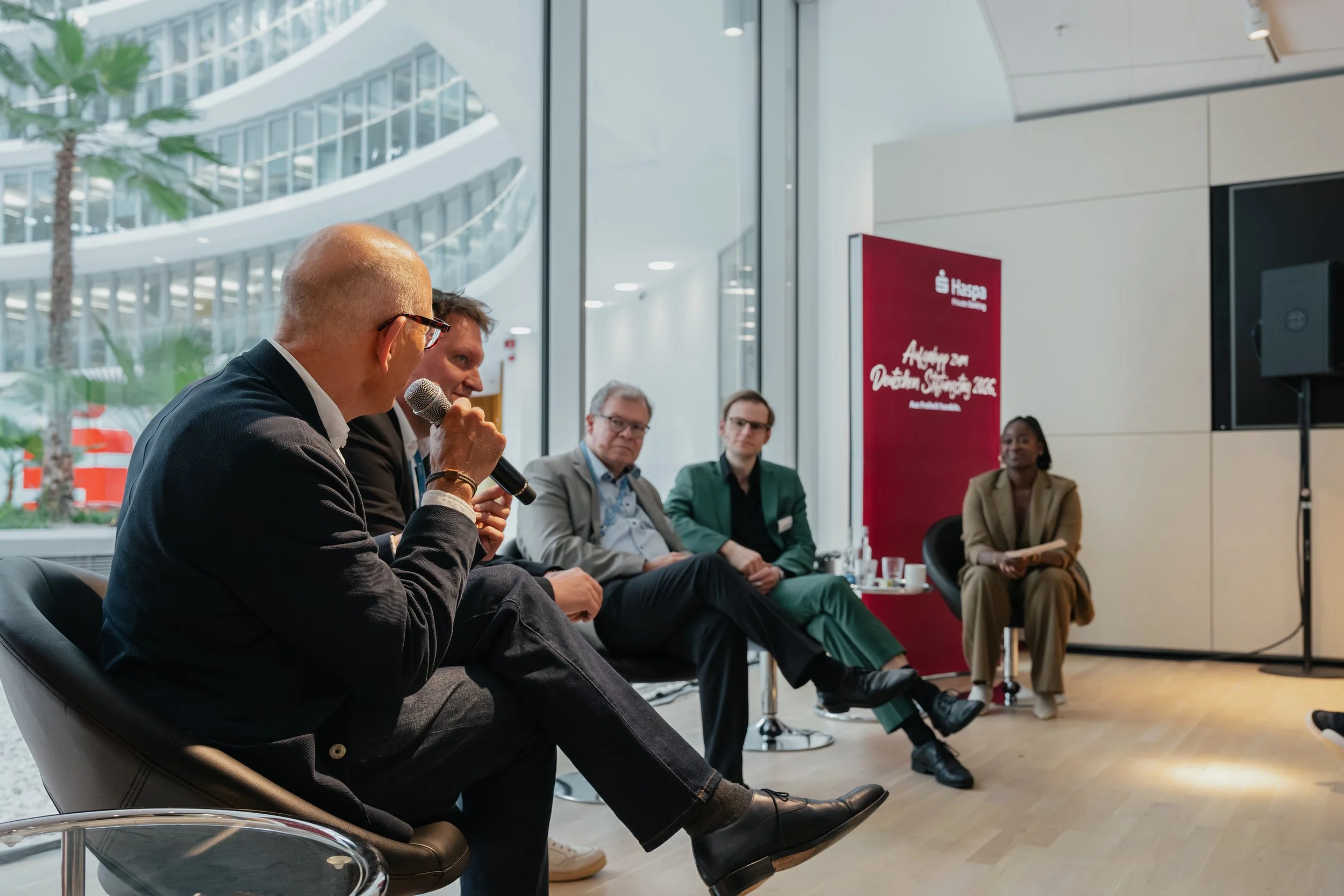 A panel of five individuals sitting in chairs during a discussion or panel event at an indoor venue with large windows and modern architecture.