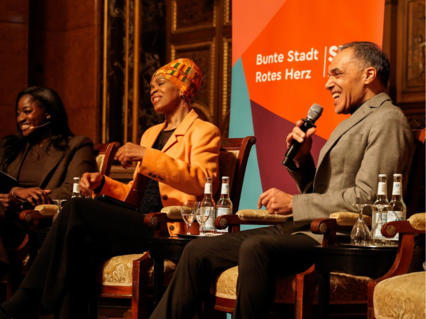 Three panelists seated on a stage during a discussion, with a colorful backdrop and microphones in hand.