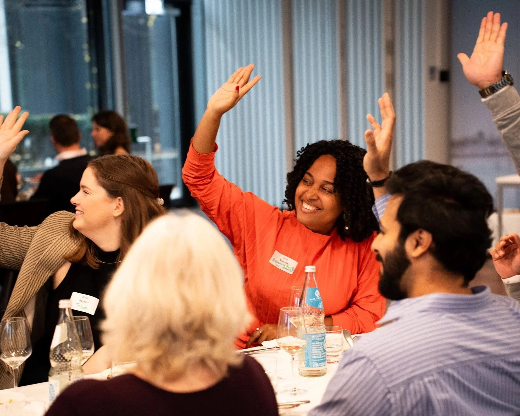 A group of people sitting around a table at a formal gathering or conference, with one woman in an orange top raising her hand and smiling.