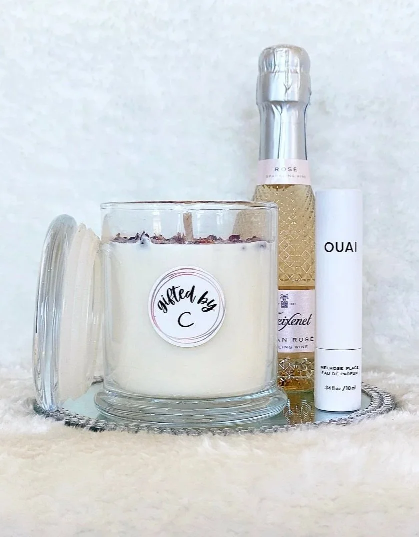 A white candle in a glass holder with a label reading "gifted by C," a bottle of Leizenet sparkling wine with a pink label, and a white cylindrical perfume or rollerball container labeled "OUAI" on a reflective tray, all on a soft, fuzzy surface.