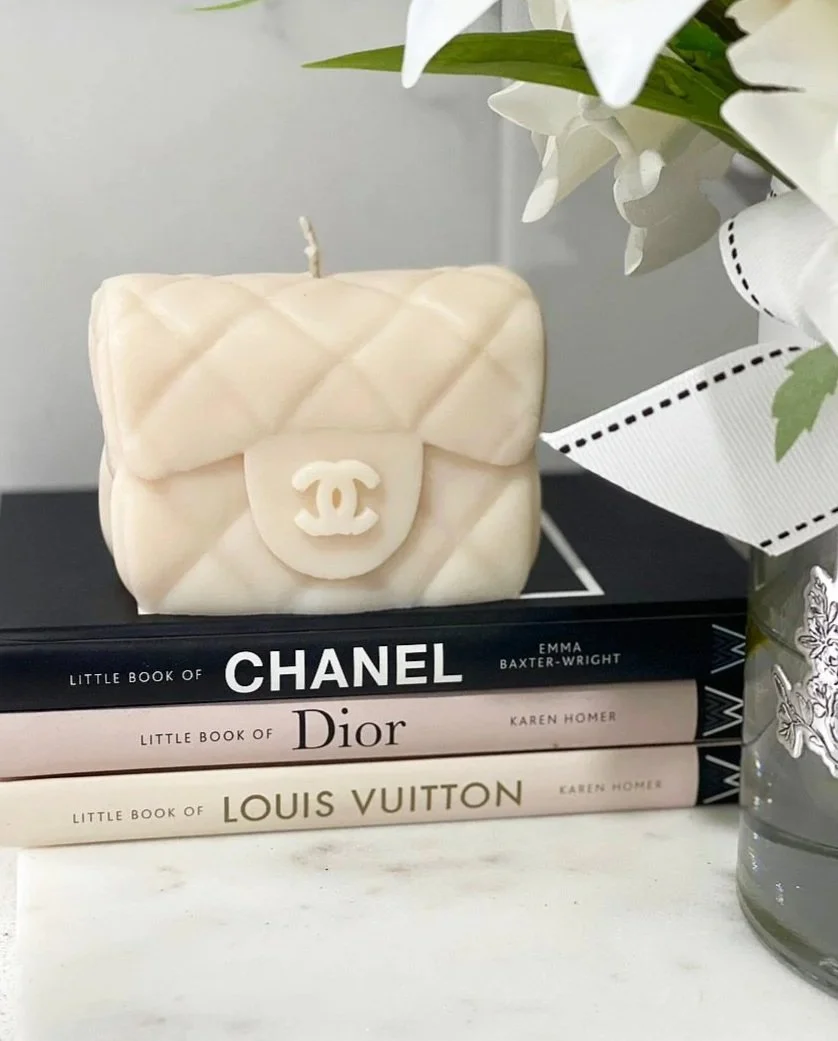 A white quilted handbag-shaped candle with the Chanel logo on the front, placed on top of three fashion books titled 'Little Book of Chanel,' 'Little Book of Dior,' and 'Little Book of Louis Vuitton,' next to a vase with white flowers and a white rib