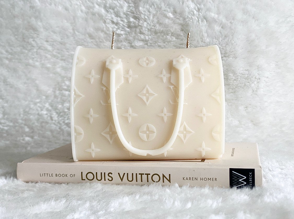 A white candle shaped like a Louis Vuitton handbag with the brand's logo pattern, placed on top of a book titled 'Little Book of Louis Vuitton' by Karen Homer, against a textured background.