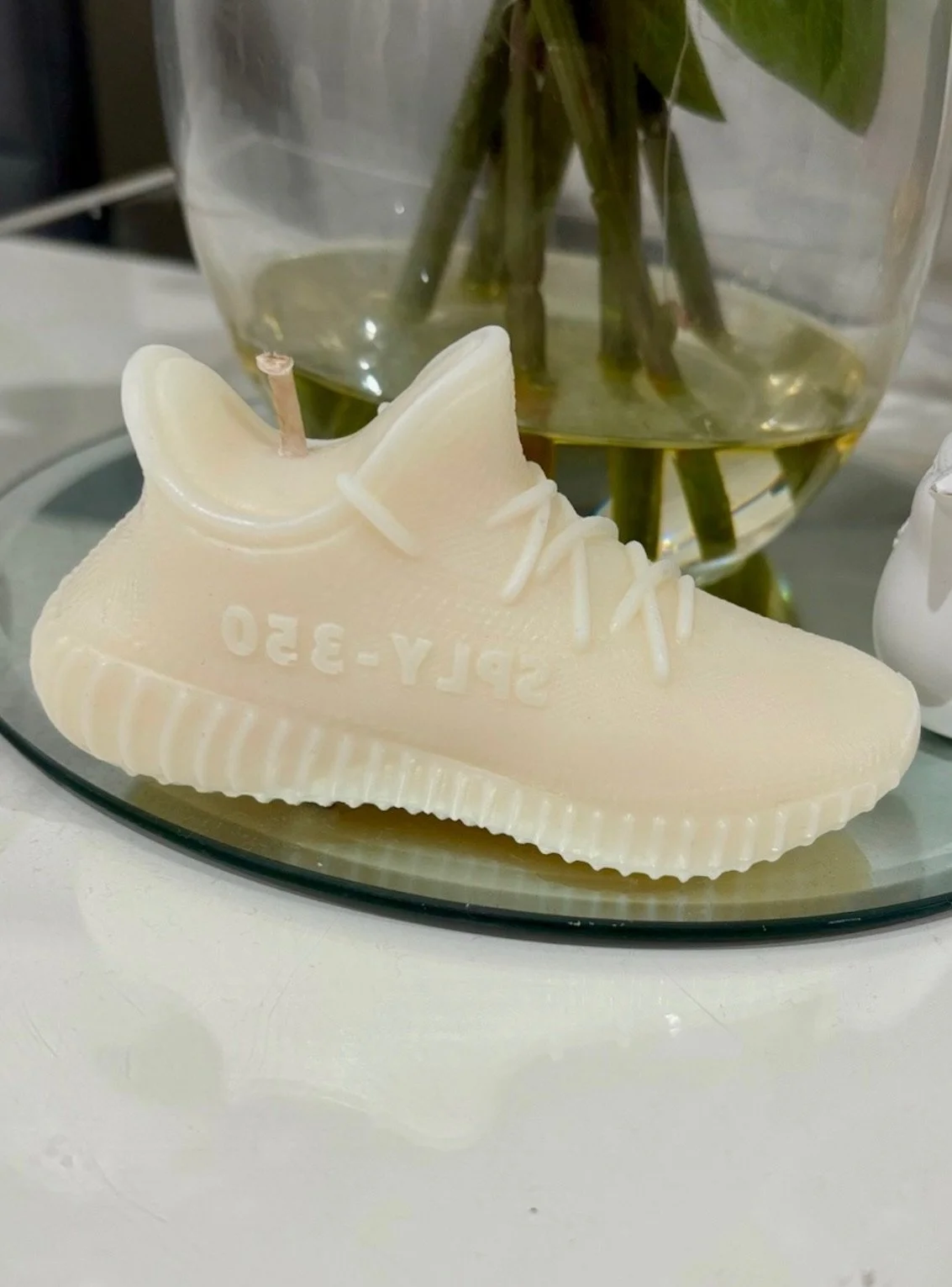 A white candle shaped like a sneaker with the inscription 'SPLY-350', placed on a tray. Behind it, there is a glass vase with water and green plant stems.
