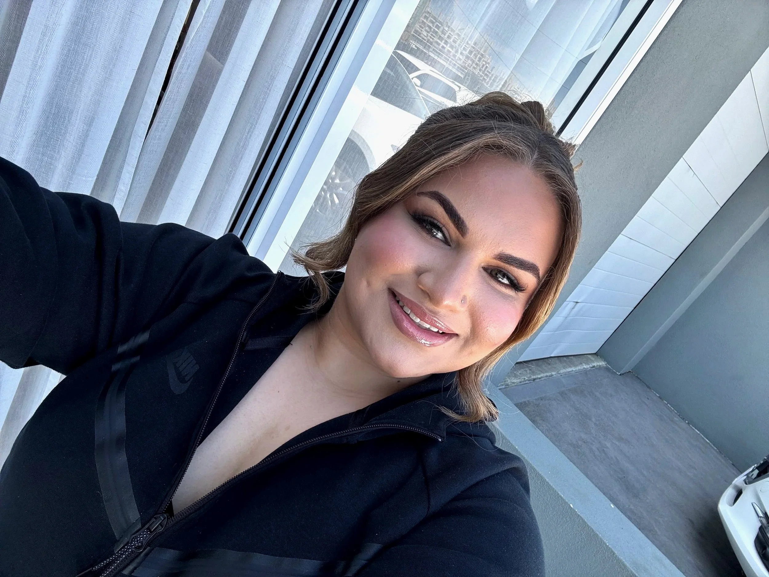 A woman with shoulder-length brown hair smiling for a selfie, wearing a black Nike jacket, standing near a large window with sheer curtains and a view of parked cars and a building outside.