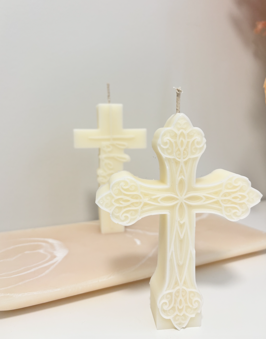 Light-colored carved candles in the shape of crosses, one in the foreground and one in the background, placed on a pinkish surface with a white swirl pattern against a plain background.
