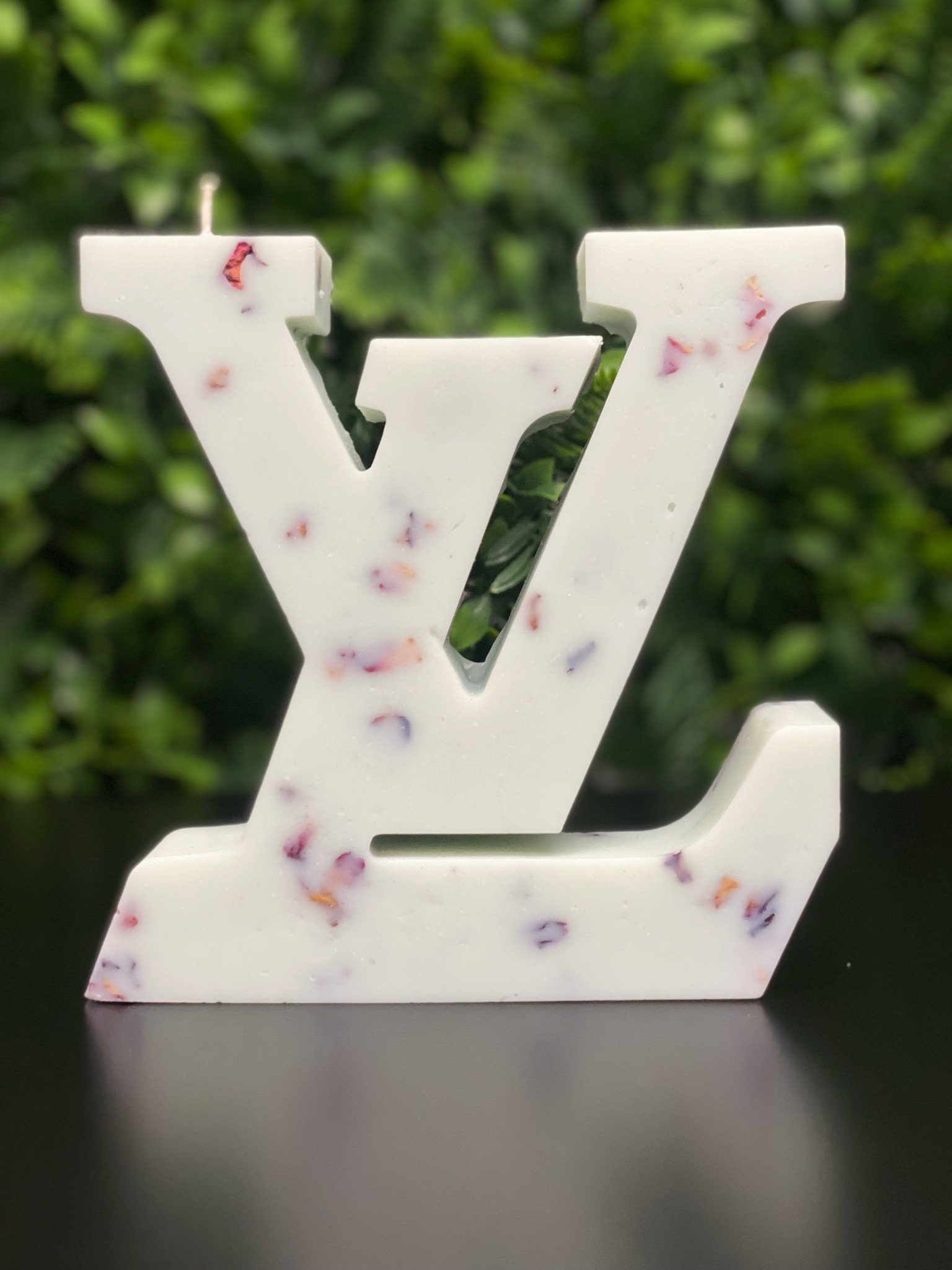 White decorative letter 'V' with small purple and red floral accents, placed on a black surface with a green leafy background.