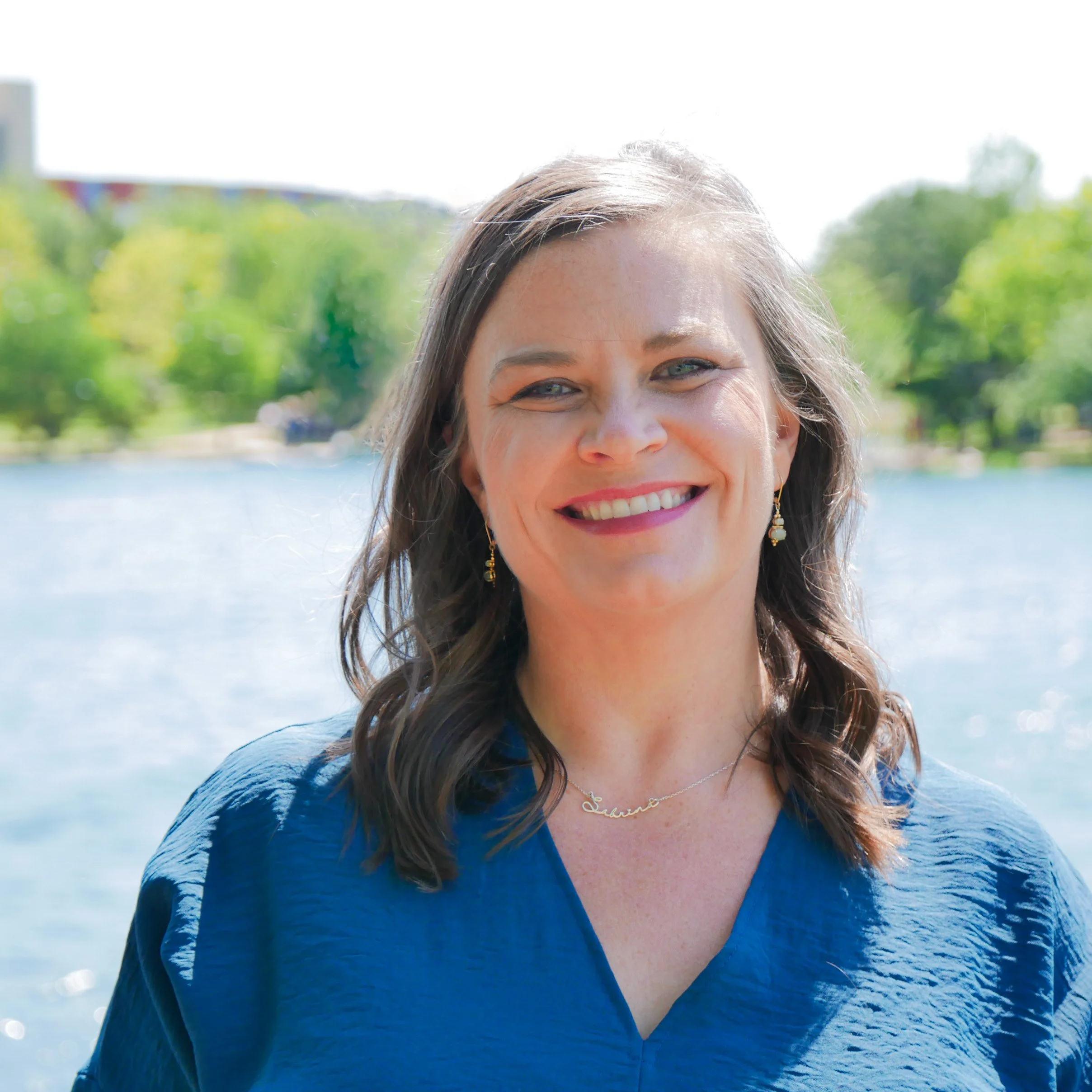 A smiling woman with shoulder-length brown hair and blue eyes standing outdoors near a lake with green trees in the background, wearing a blue top, earrings, and a necklace.