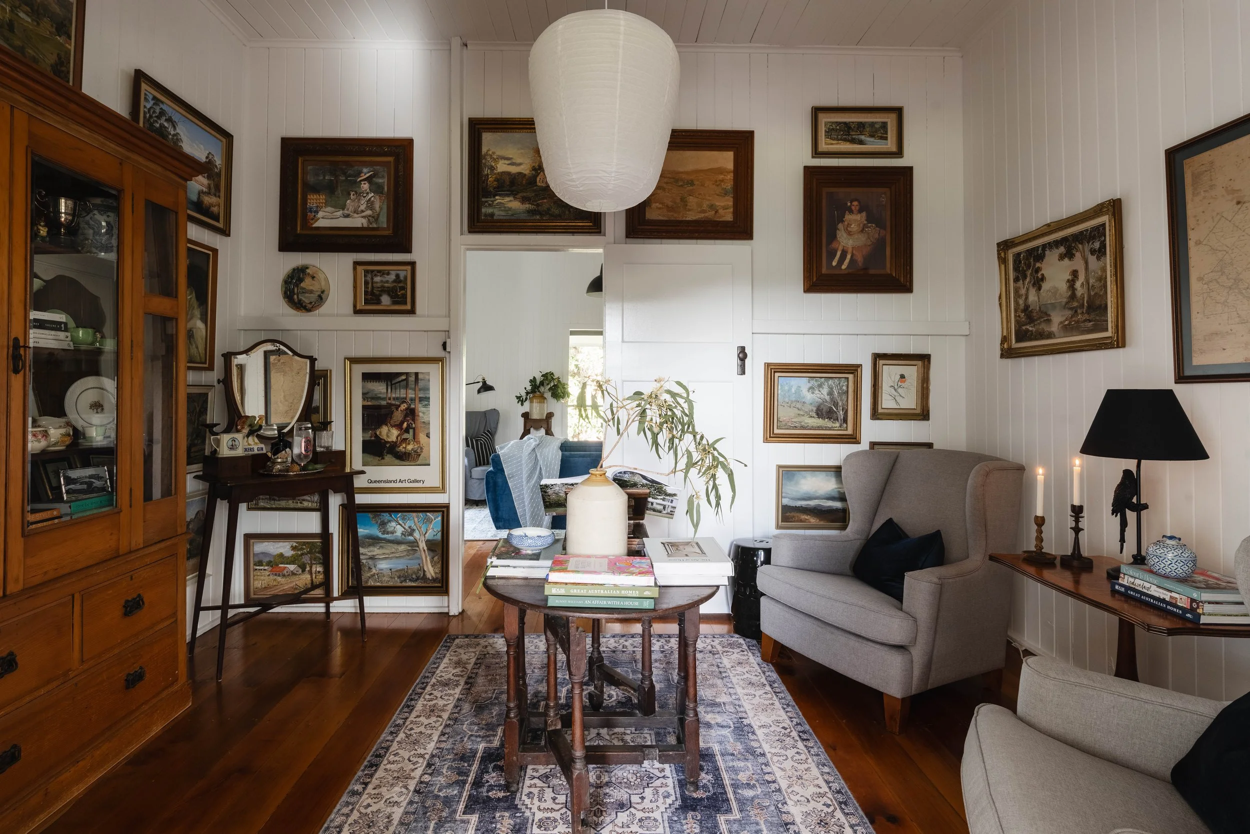 A cozy living room with white paneled walls, decorated with numerous framed paintings and pictures. There is a large wooden cabinet on the left, a central wooden table with books and a large white vase, a gray armchair, and a side table with a lamp and books. The room features a patterned rug on a wooden floor, with an open doorway leading to a dining area with a blue upholstered chair visible in the background.