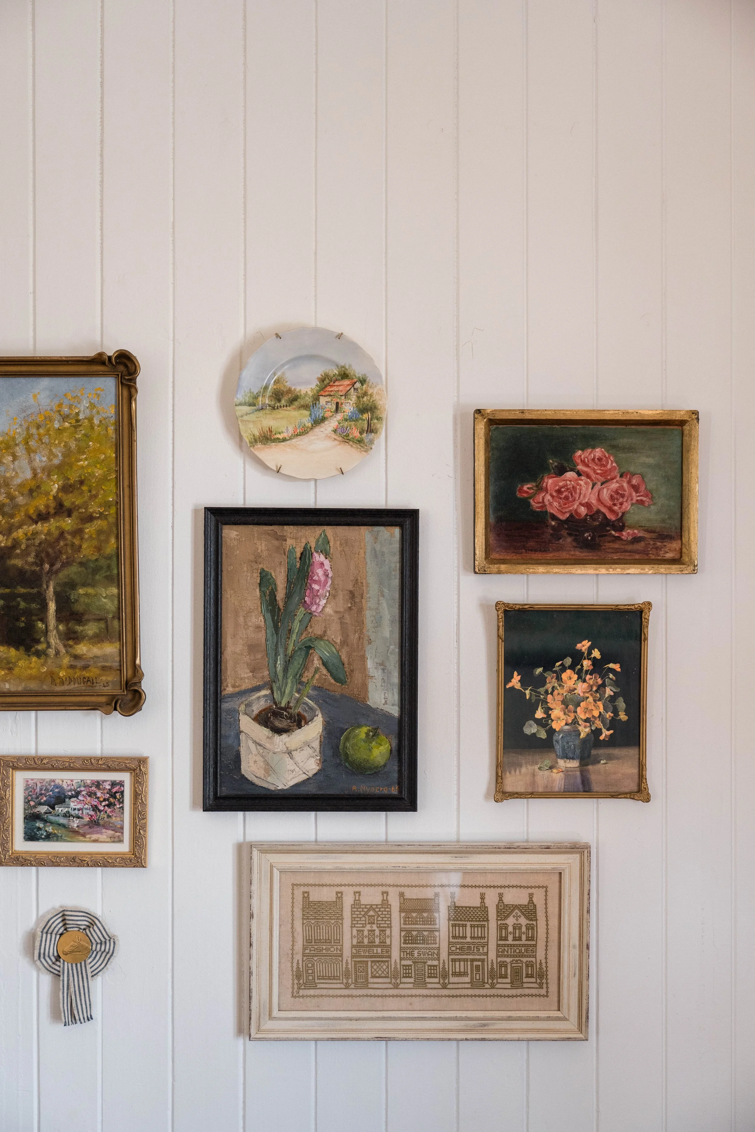 A collection of seven framed artworks and decorative items on a white paneled wall, including paintings of a tree, a house, flowers, a potted flower, and a cross-stitched pattern, with a ribbon and medal below.