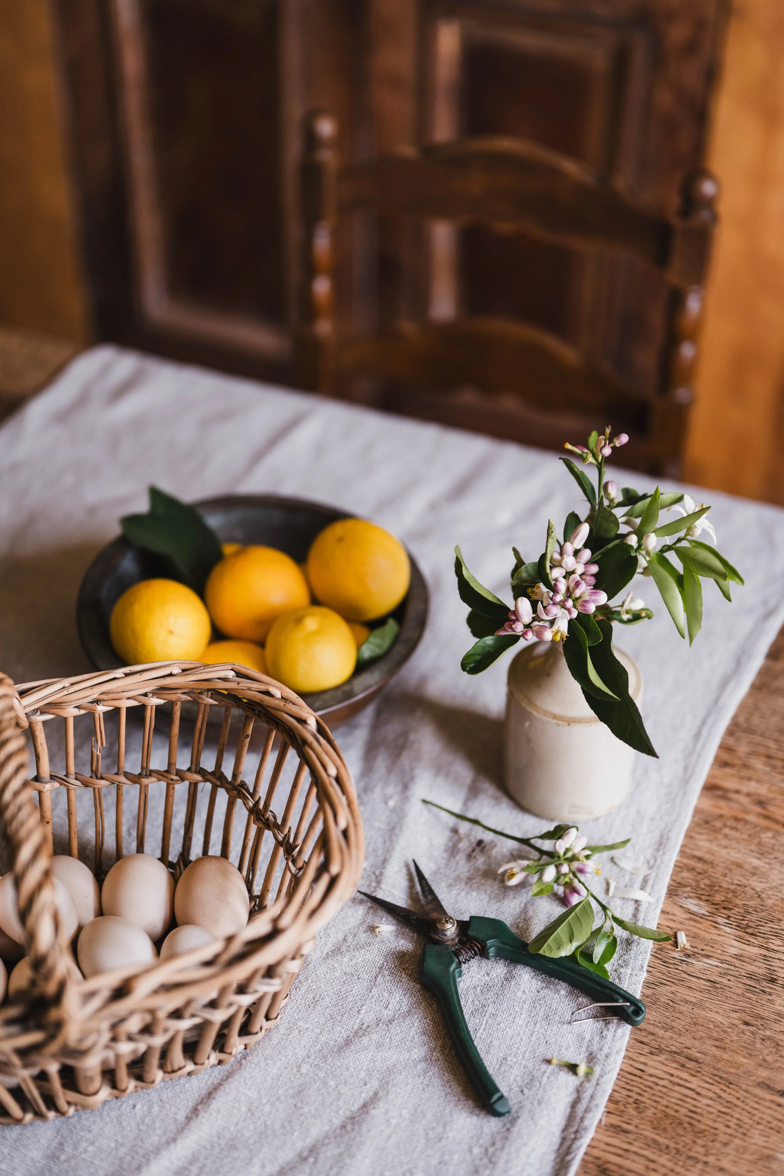 A rustic table decorated with a white linen cloth, holding a woven basket of white eggs, a dark bowl of lemons with leaves, a small vase with pink and white flowers, and gardening shears.