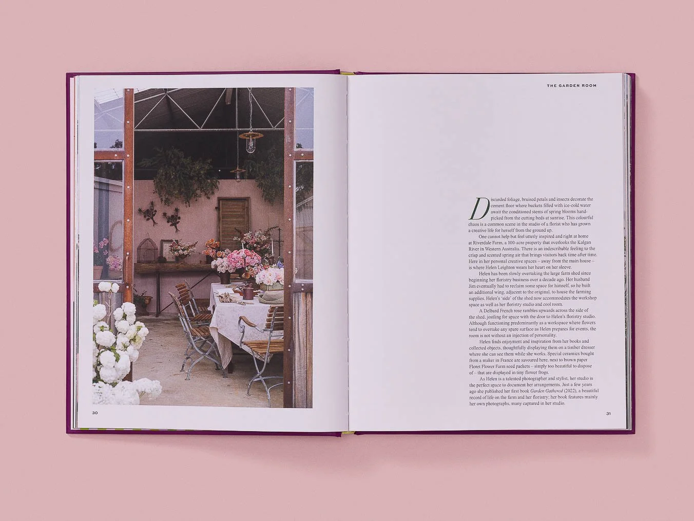 Open book on pink surface, showing a photo of a greenhouse or garden room with a dining table, chairs, and floral arrangements.