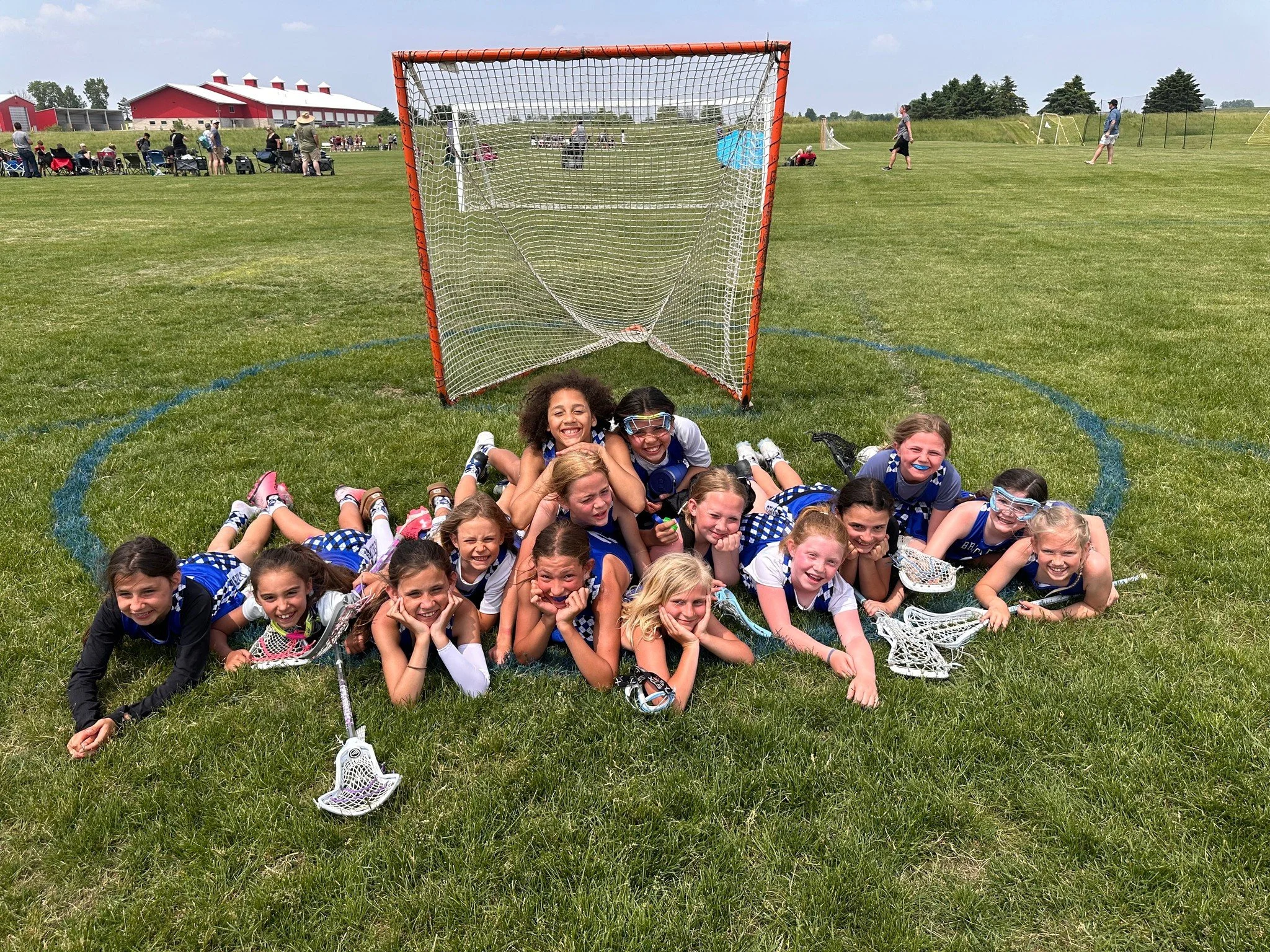A group of young girls in matching blue and white sports uniforms are lying on the grass in front of a lacrosse goal, smiling and posing for a photo, on a sunny day at a sports field.