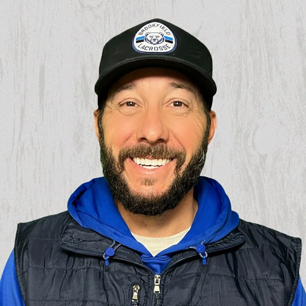 Smiling man with beard wearing a black cap with a letter B and crossed lacrosse sticks, blue hoodie, and black vest, standing against a light textured background.