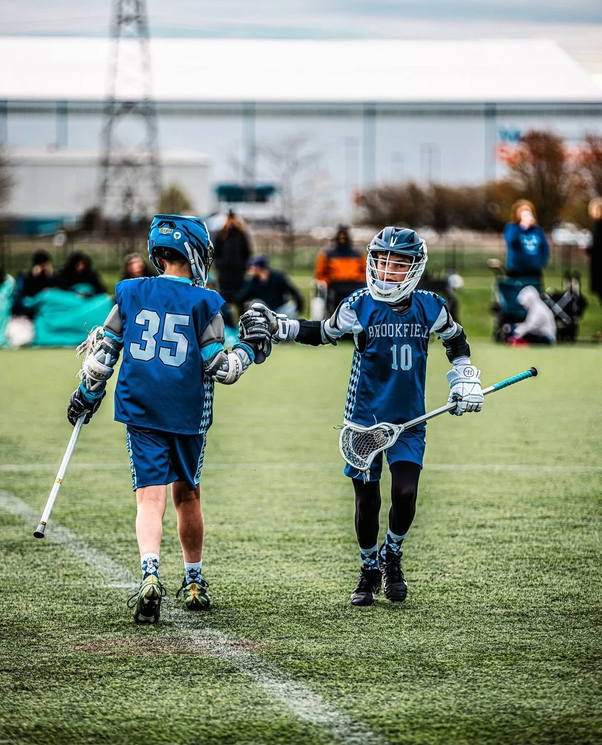 Two young boys in lacrosse uniforms shake hands on a field during a game. One is wearing jersey number 35, and the other number 10. Both are dressed in blue uniforms with helmets and carrying lacrosse sticks.