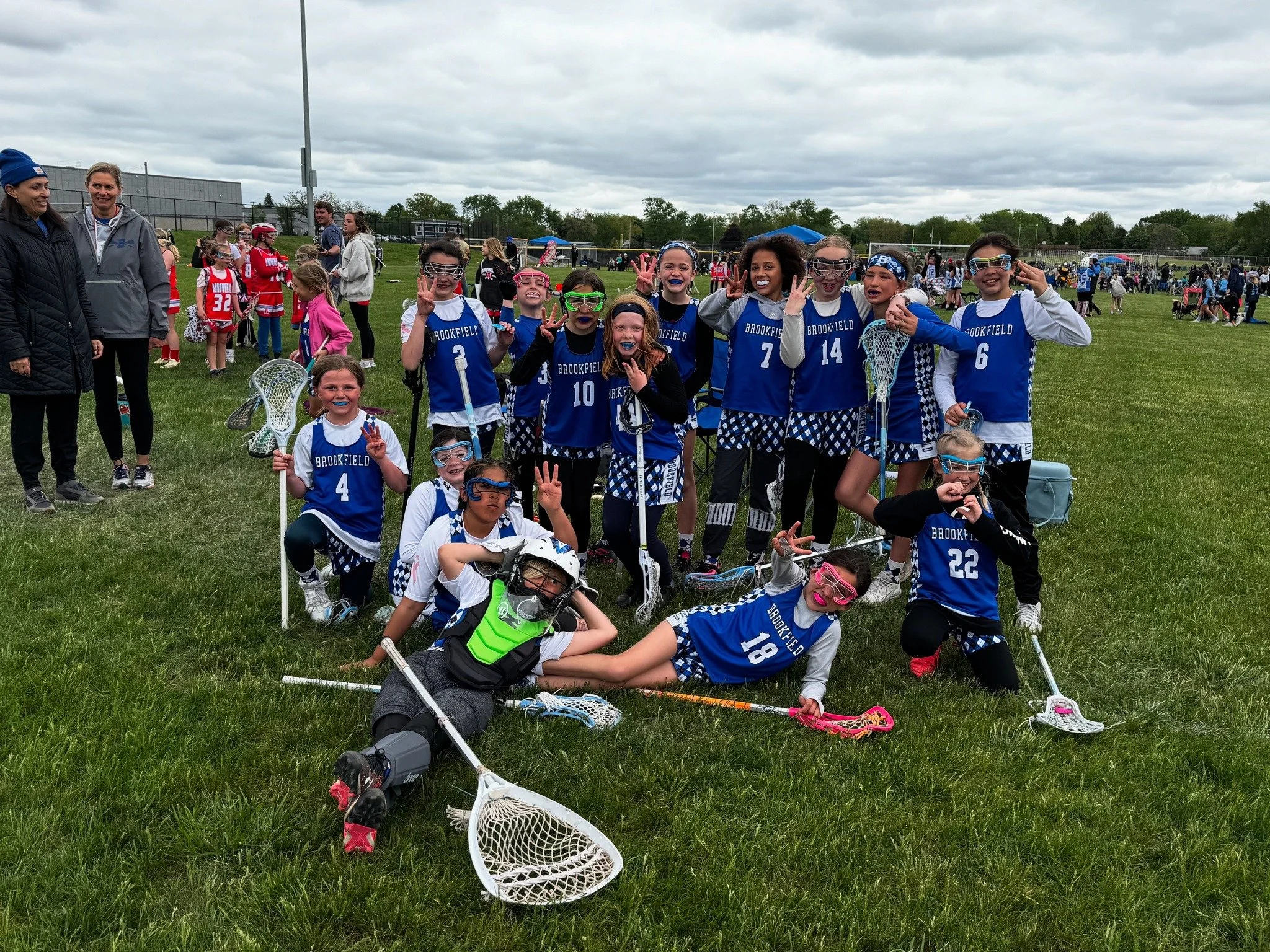 Group of young girls in blue lacrosse uniforms posing together on a grassy field during a sports event with spectators in the background.