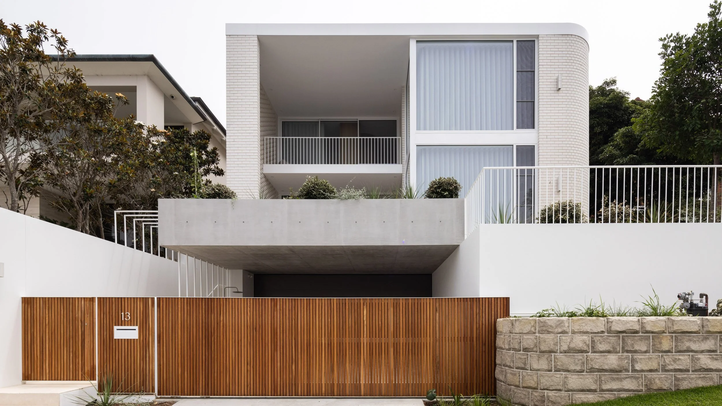 Modern white multi-story house with balcony, white railing, and wooden gate, surrounded by greenery and stone wall.