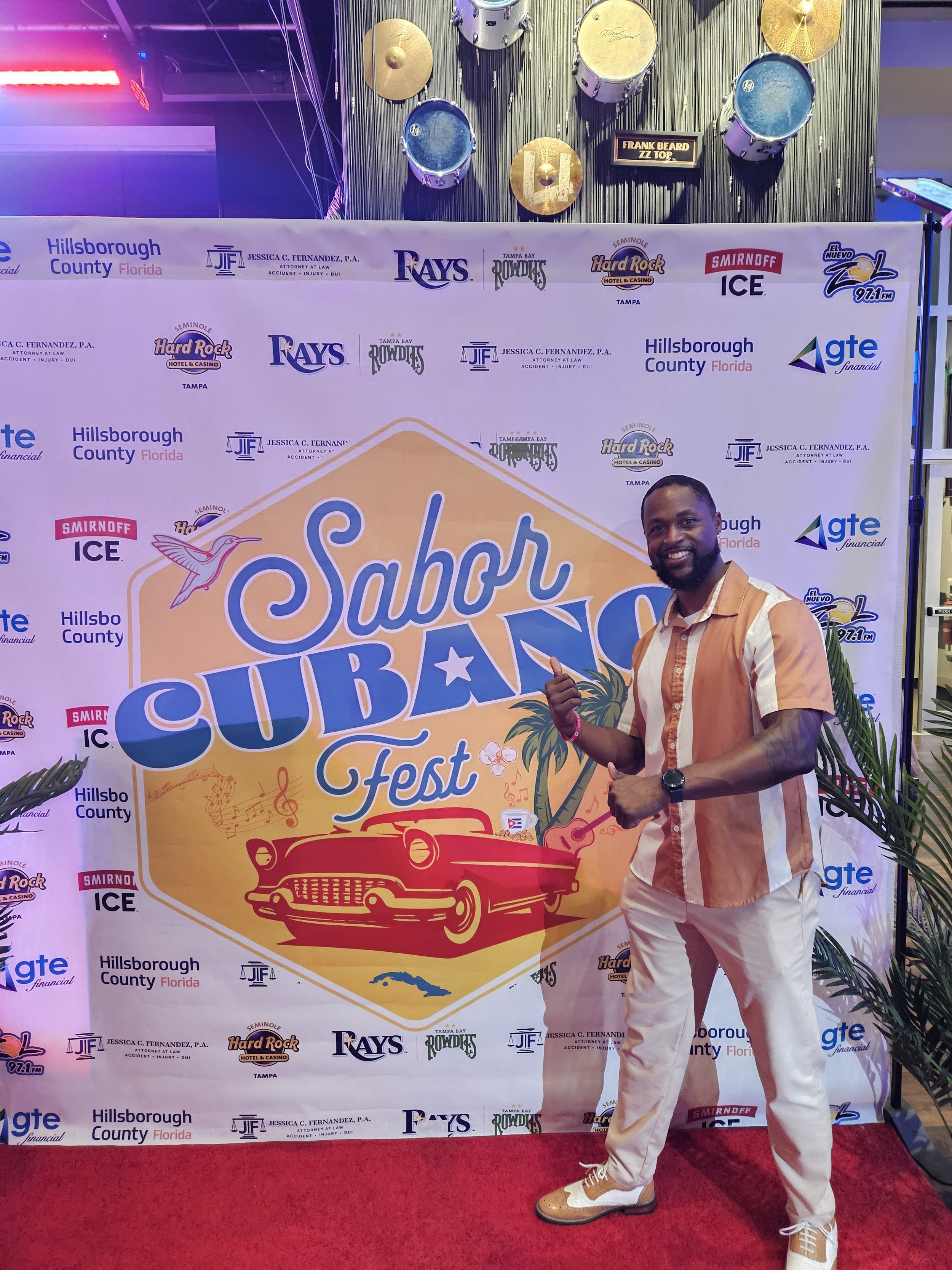 A man posing in front of a backdrop at the Sabor Cubano Fest, decorated with logos and a large graphic of a classic car in Havana style. The backdrop includes logos from sponsors like Hillsborough County, Seminole Hard Rock, and more. A set of drums 