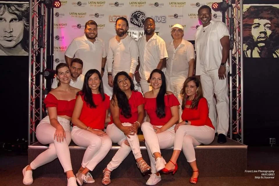 Group of people posing at a Latin-themed event. The men are wearing white, and the women are in red tops with white pants. They're smiling and standing against a backdrop with 'Latin Night' repeated on it. There are portraits of famous musicians on t
