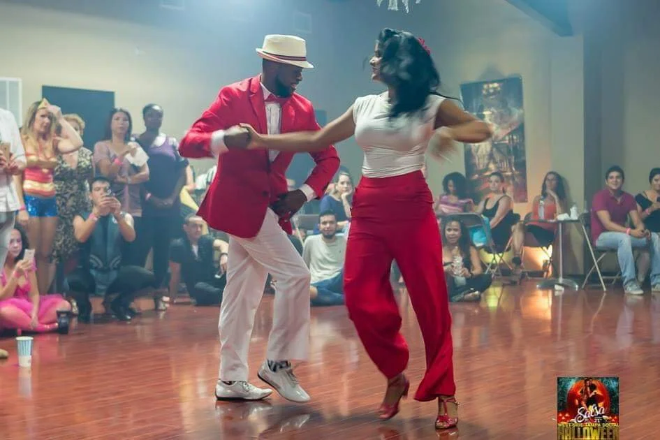 Dancers in a lively event, with a man in a red jacket and white pants, wearing a hat, and a woman in red pants and a white shirt, performing in front of a seated audience in a room with polished wooden floors.