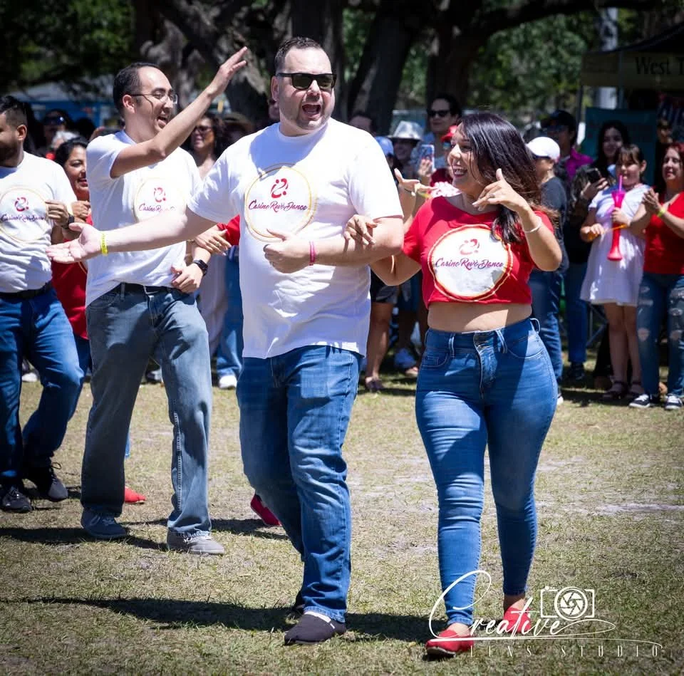 People dancing outdoors at a festival, wearing casual clothes like jeans and t-shirts, with others in the background cheering and watching.