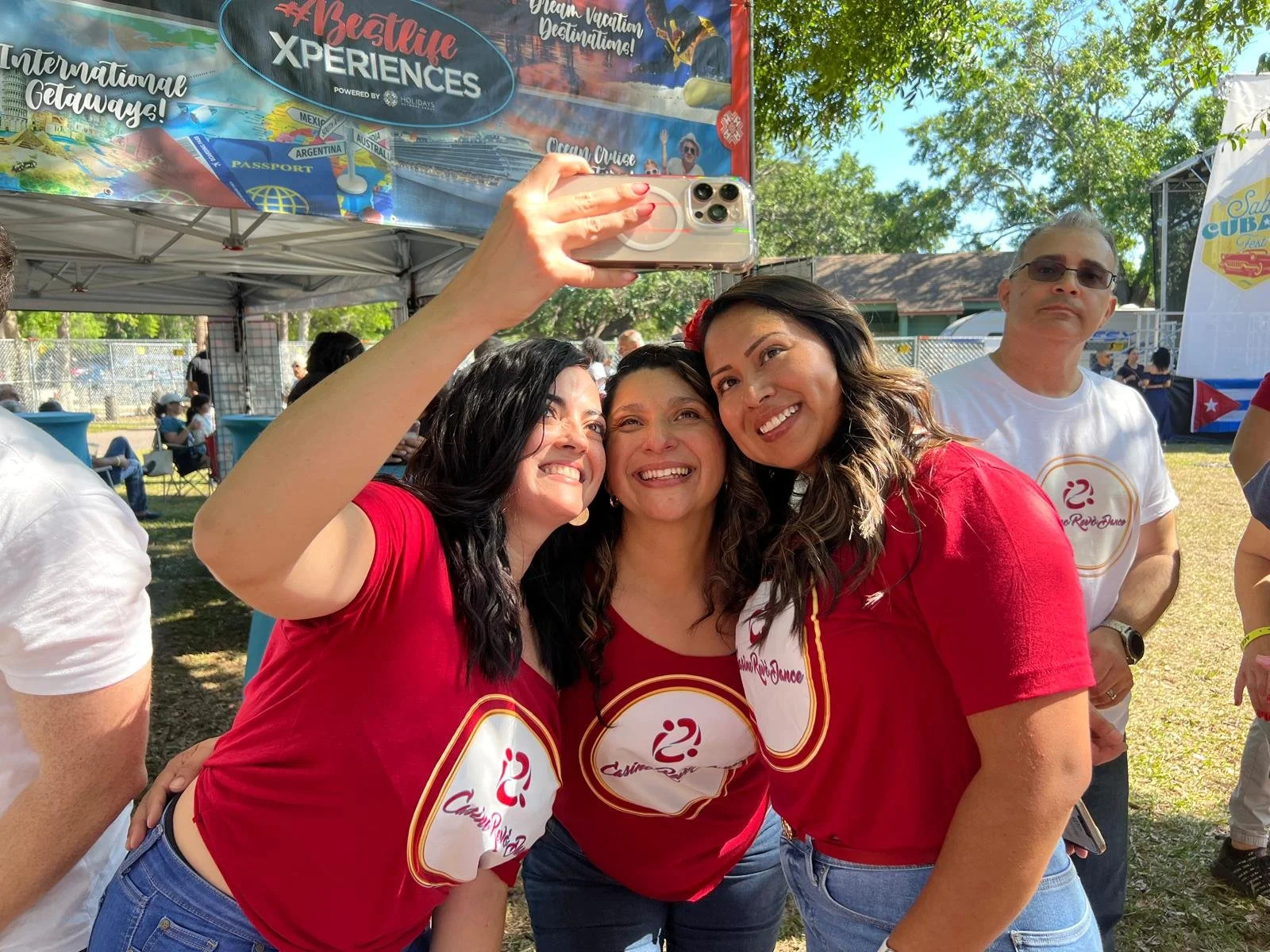 Three women in red shirts taking a selfie at an outdoor event, with a man standing in the background. A tent and promotional banners are visible.