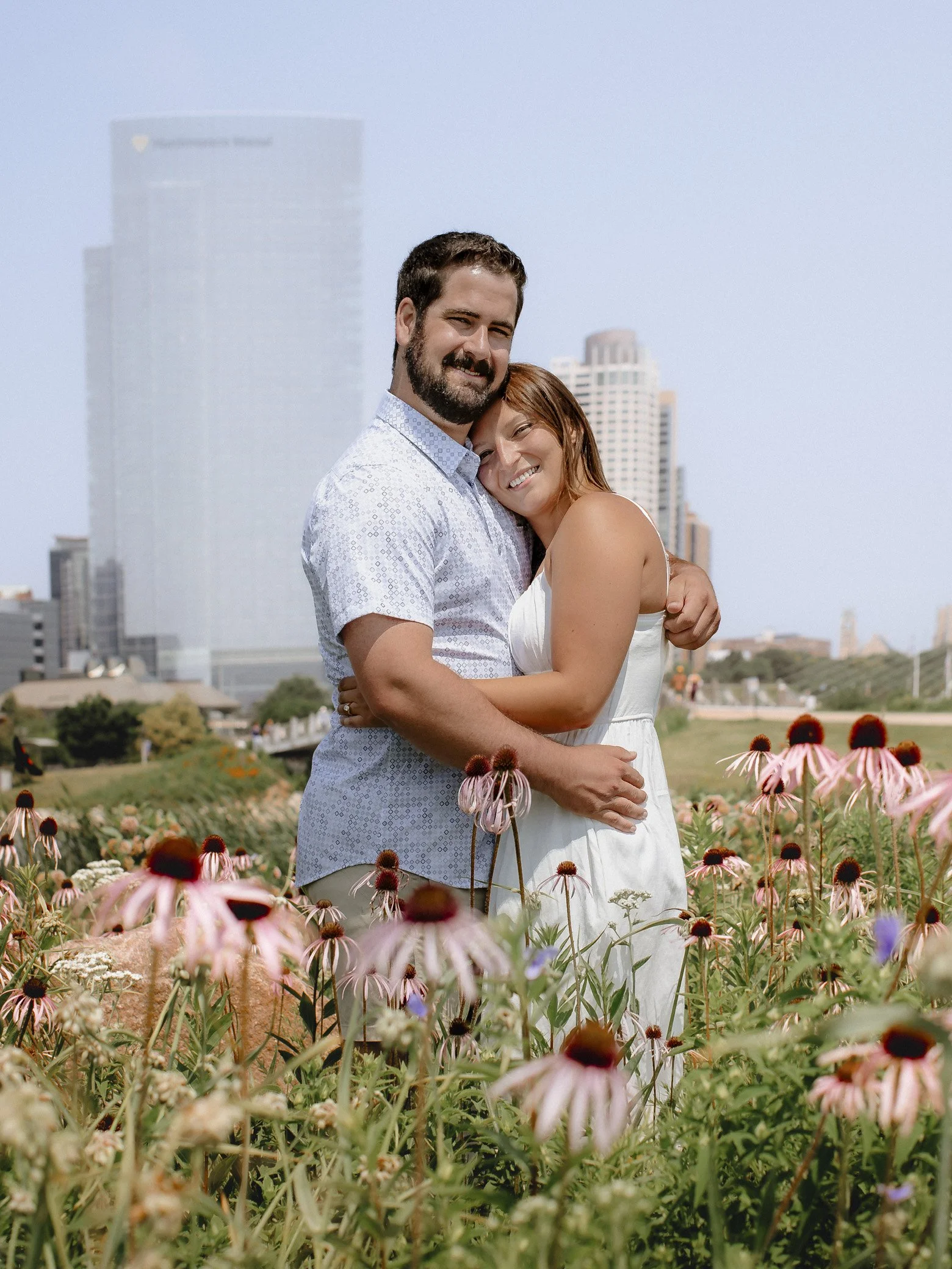 A couple smiling and hugging in a flower field with city buildings in the background.