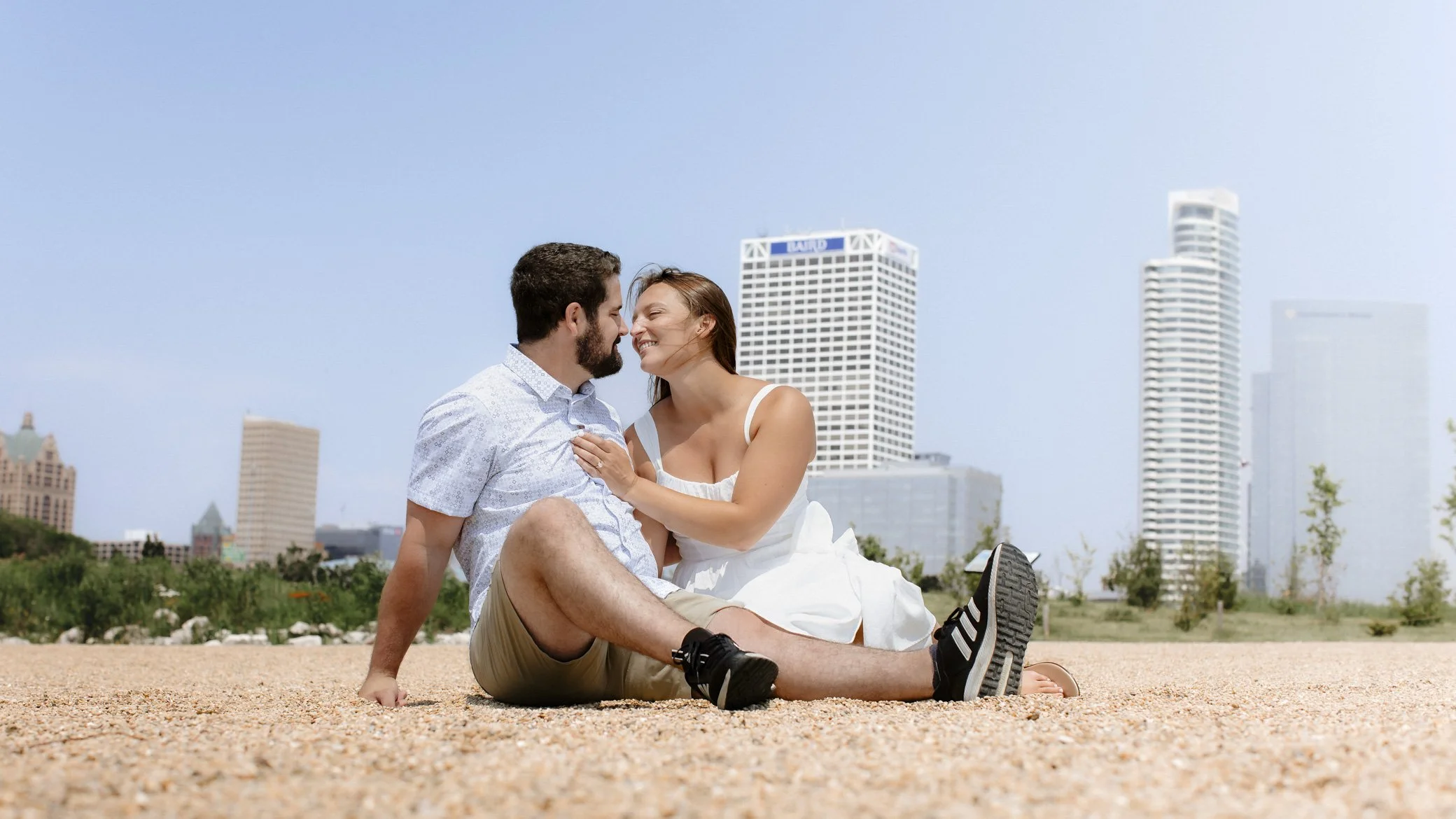 A young couple sitting on gravel ground outdoors, smiling and enjoying each other's company with a city skyline in the background.