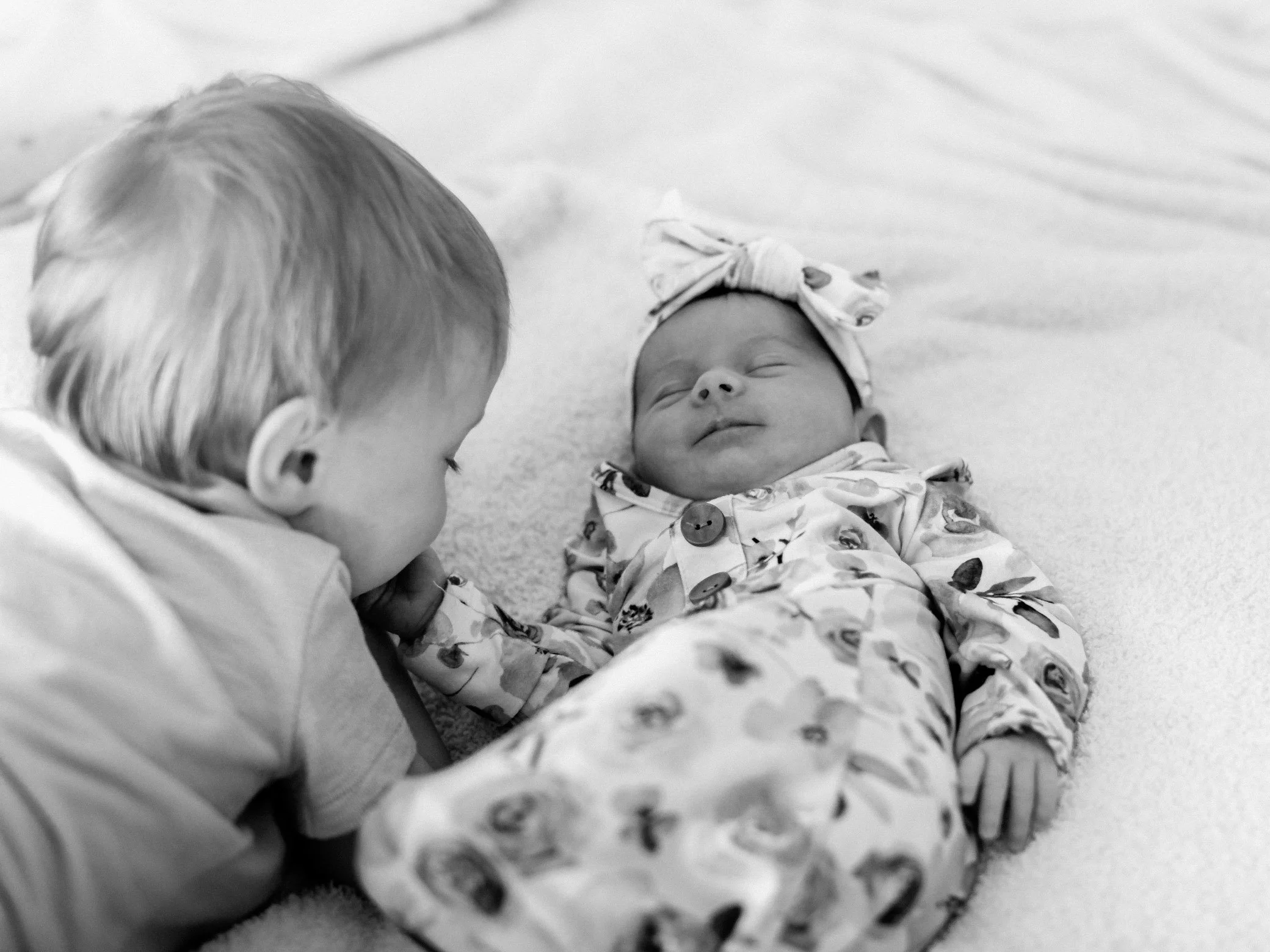 A young child with light hair looking at a sleeping baby girl dressed in a patterned onesie with a matching bow headband on a soft surface.