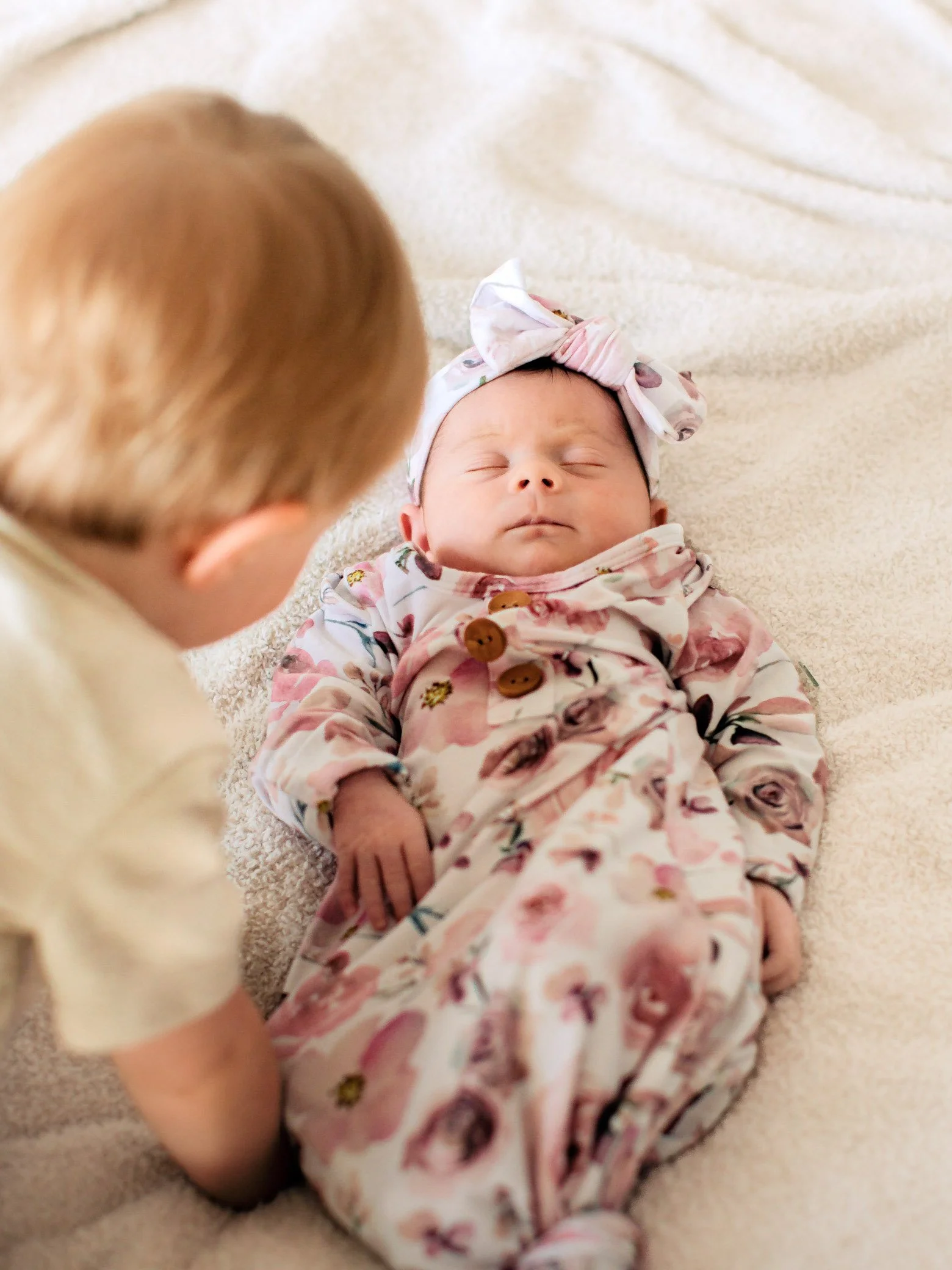 A toddler with red hair leaning over a sleeping newborn girl on a soft, cream-colored blanket. The baby is dressed in floral pajamas with a matching headband.