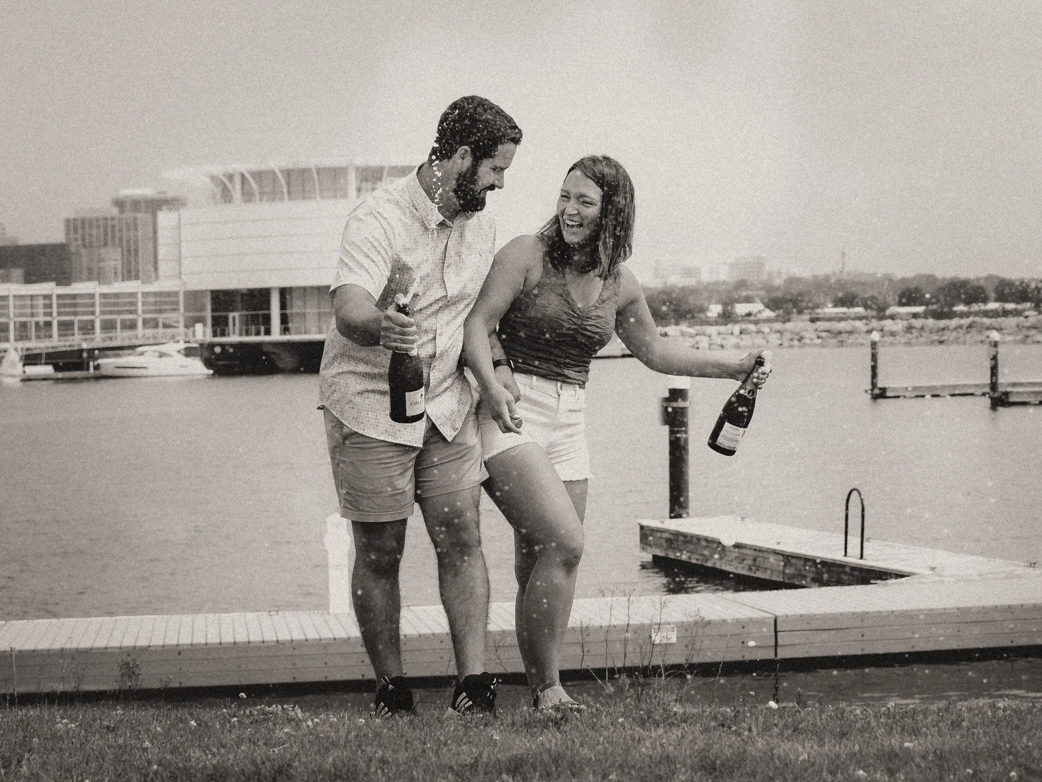 A black and white photo of a man and woman celebrating by a lake, holding bottles of champagne, with a dock and modern building in the background.