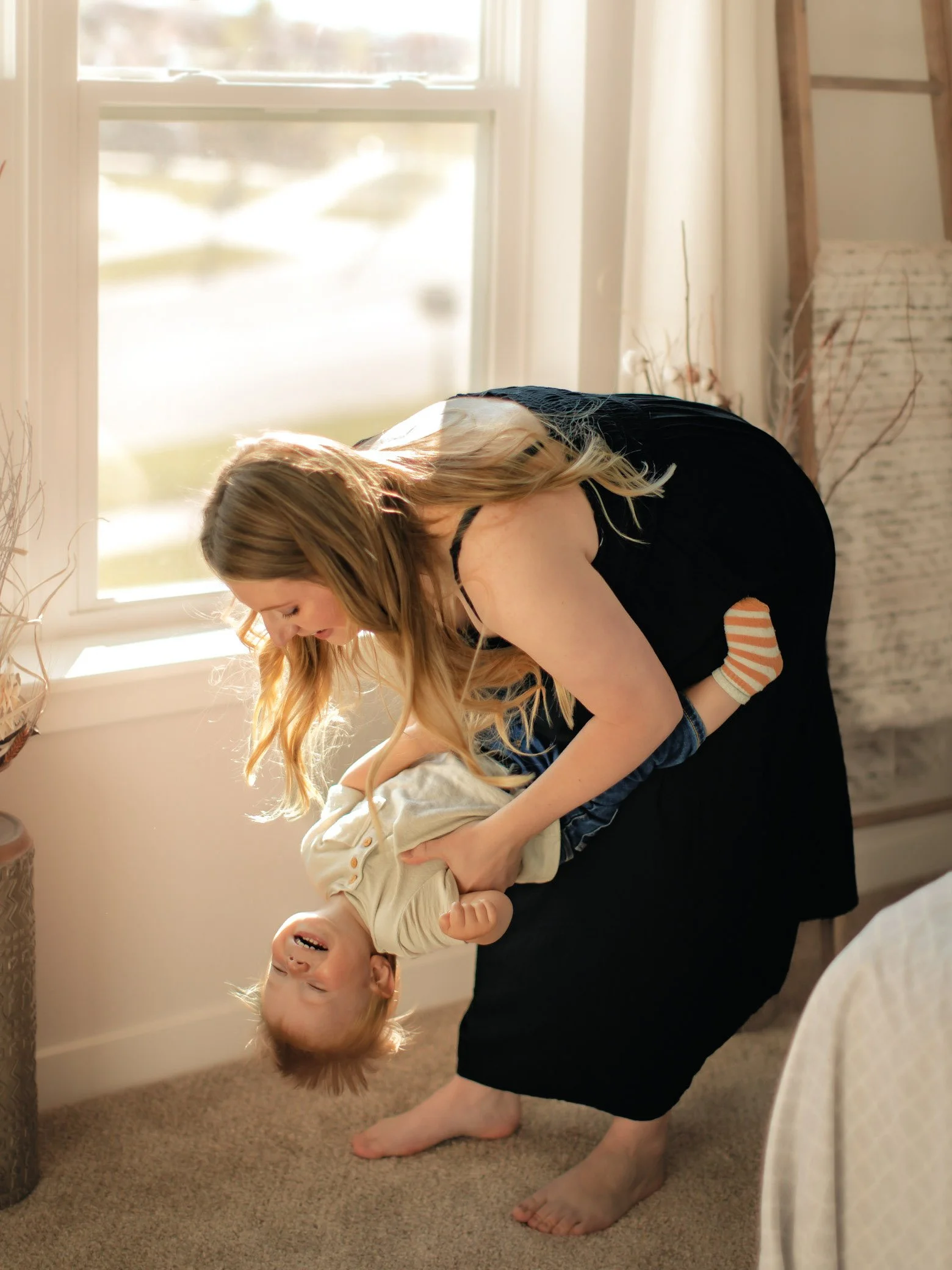 A woman playing with a young child, hanging upside down, in a brightly lit room near a window.