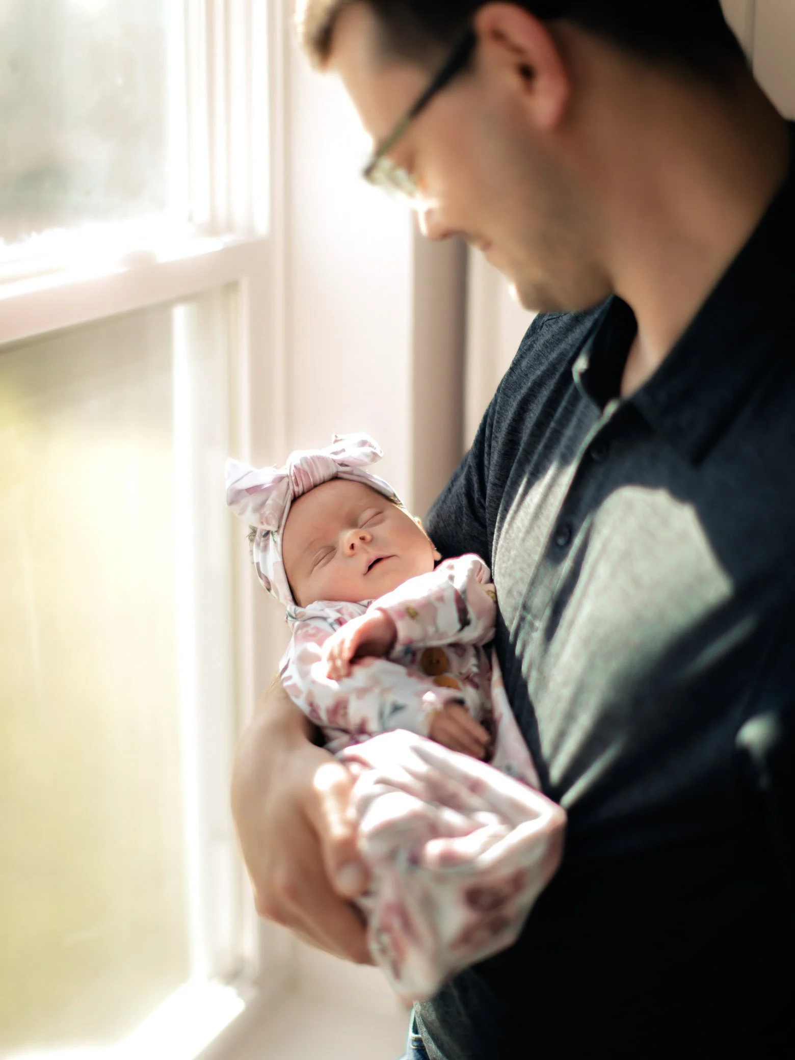 A man holding a sleeping baby girl near a window, with sunlight illuminating the scene. The girl is dressed in a floral outfit with a matching headband.