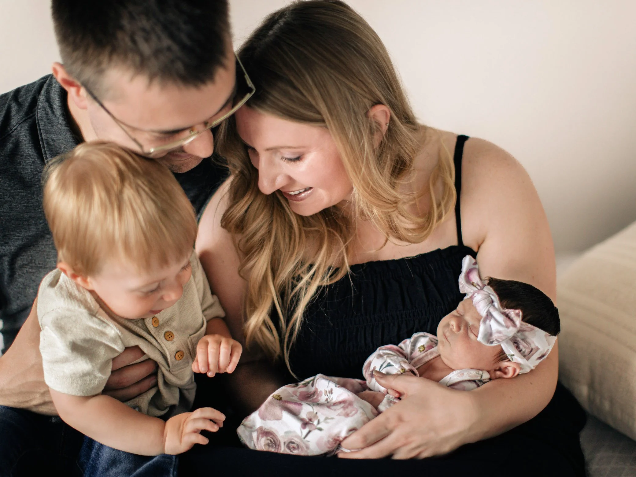 A family of four, including two children and two adults, gathered around and smiling at a newborn baby girl. The mother is holding the baby, who is dressed in a floral outfit and headband. The older boy leans in, and the father also looks on with aff