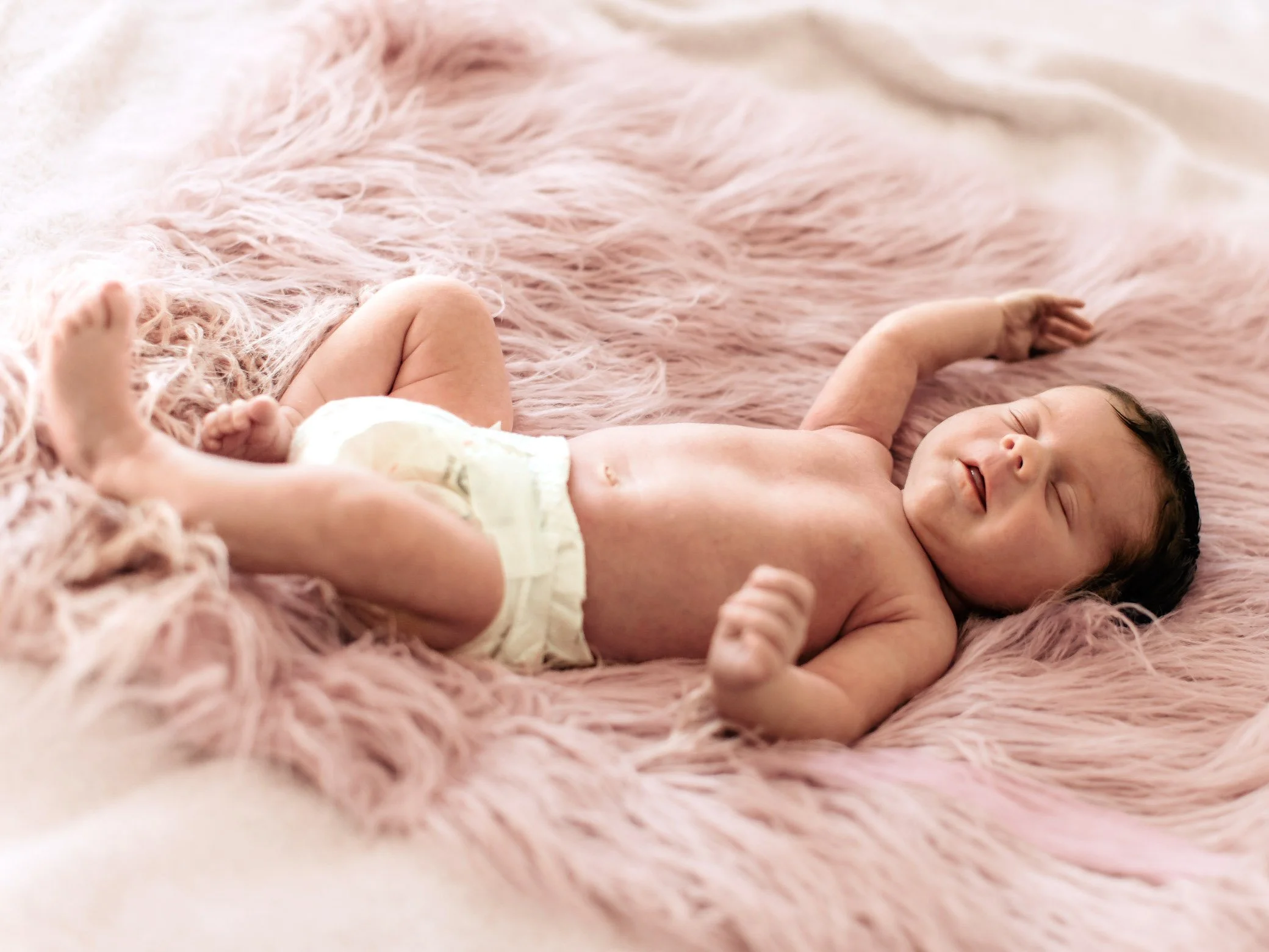 A newborn baby sleeping on a soft pink fluffy blanket wearing a diaper.