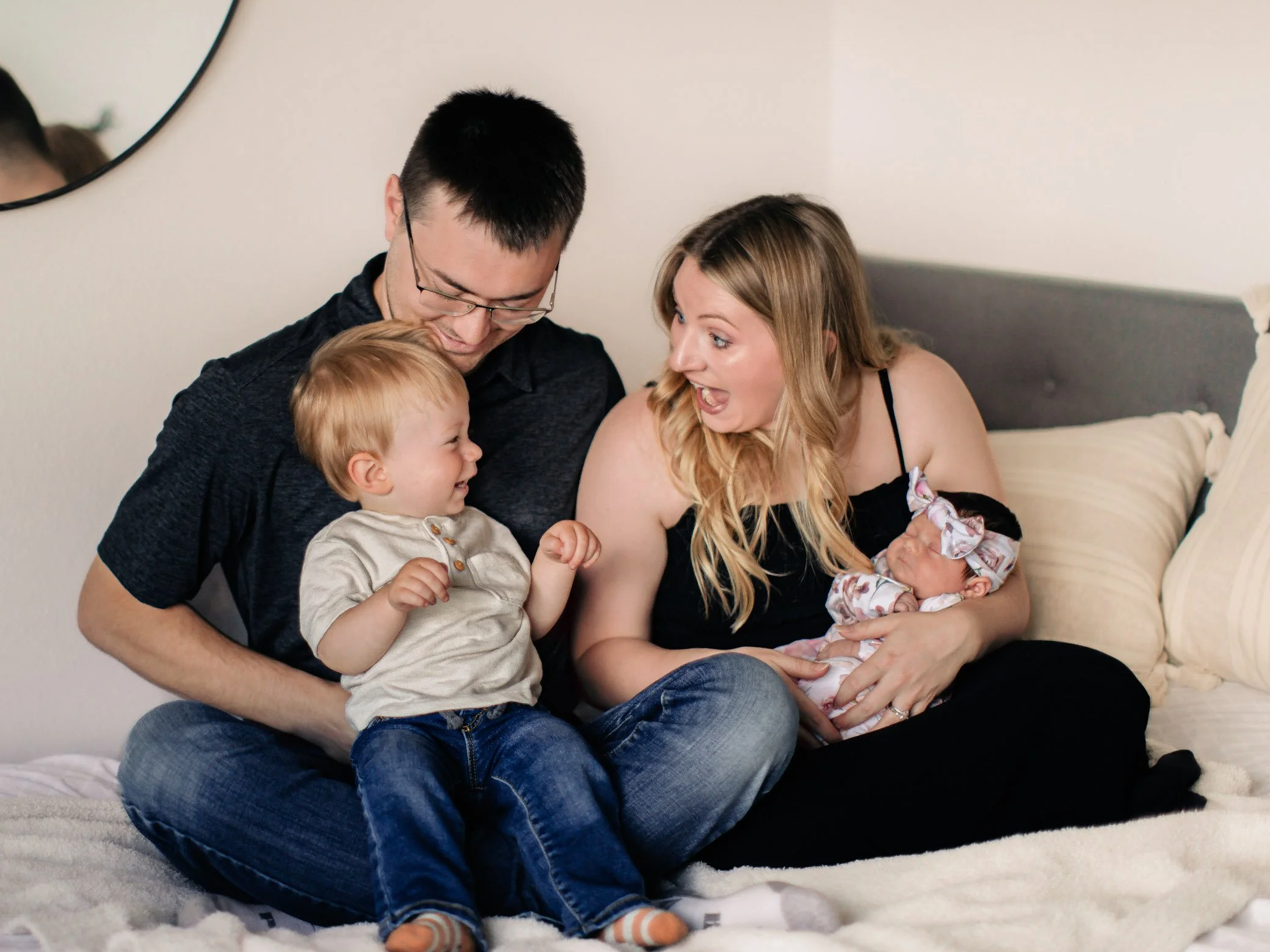 A family sitting on a bed, celebrating the birth of a newborn girl with a joyful mother, father, young son, and a baby girl dressed in a floral headband.