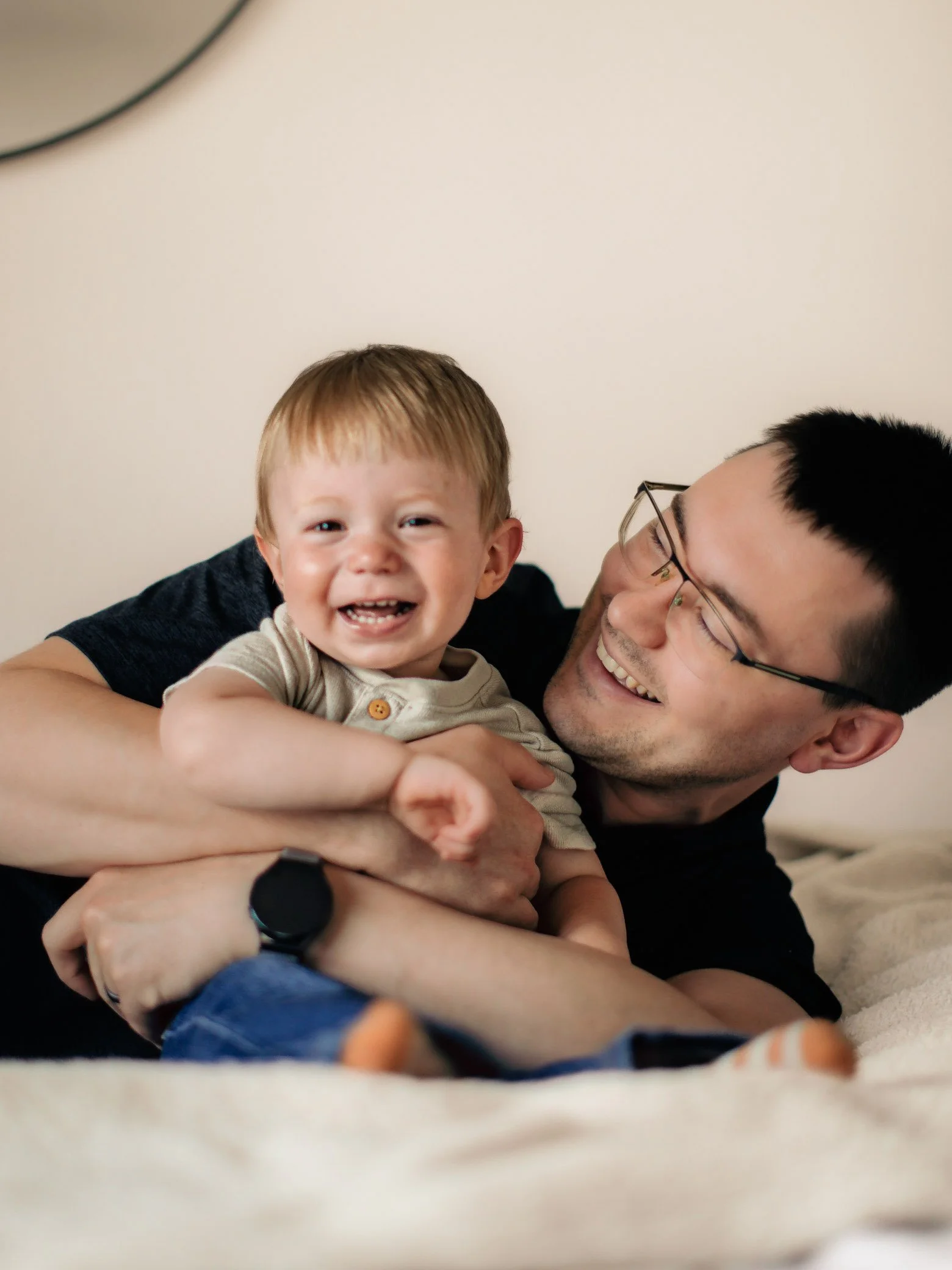 A man with glasses and a smiling face holding a young boy with a happy expression, lying on a bed in a cozy indoor setting.