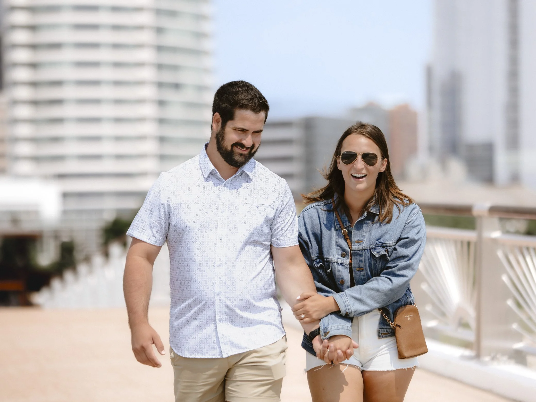 A smiling man and woman walking outdoors in an urban setting, holding hands and enjoying each other's company on a sunny day.