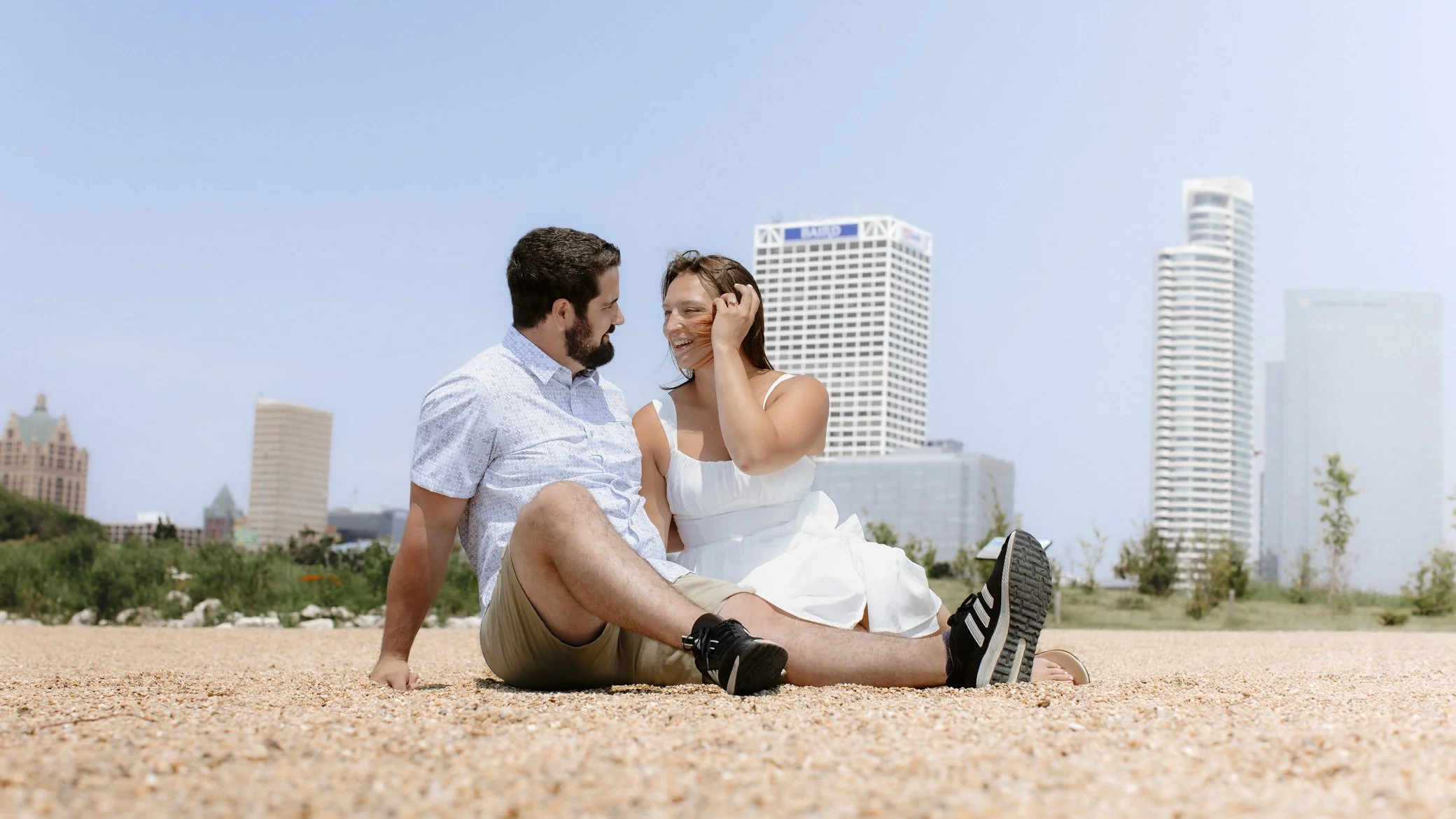 A young couple sitting on a sandy surface in an urban park, looking at each other and smiling, with tall buildings and clear sky in the background.