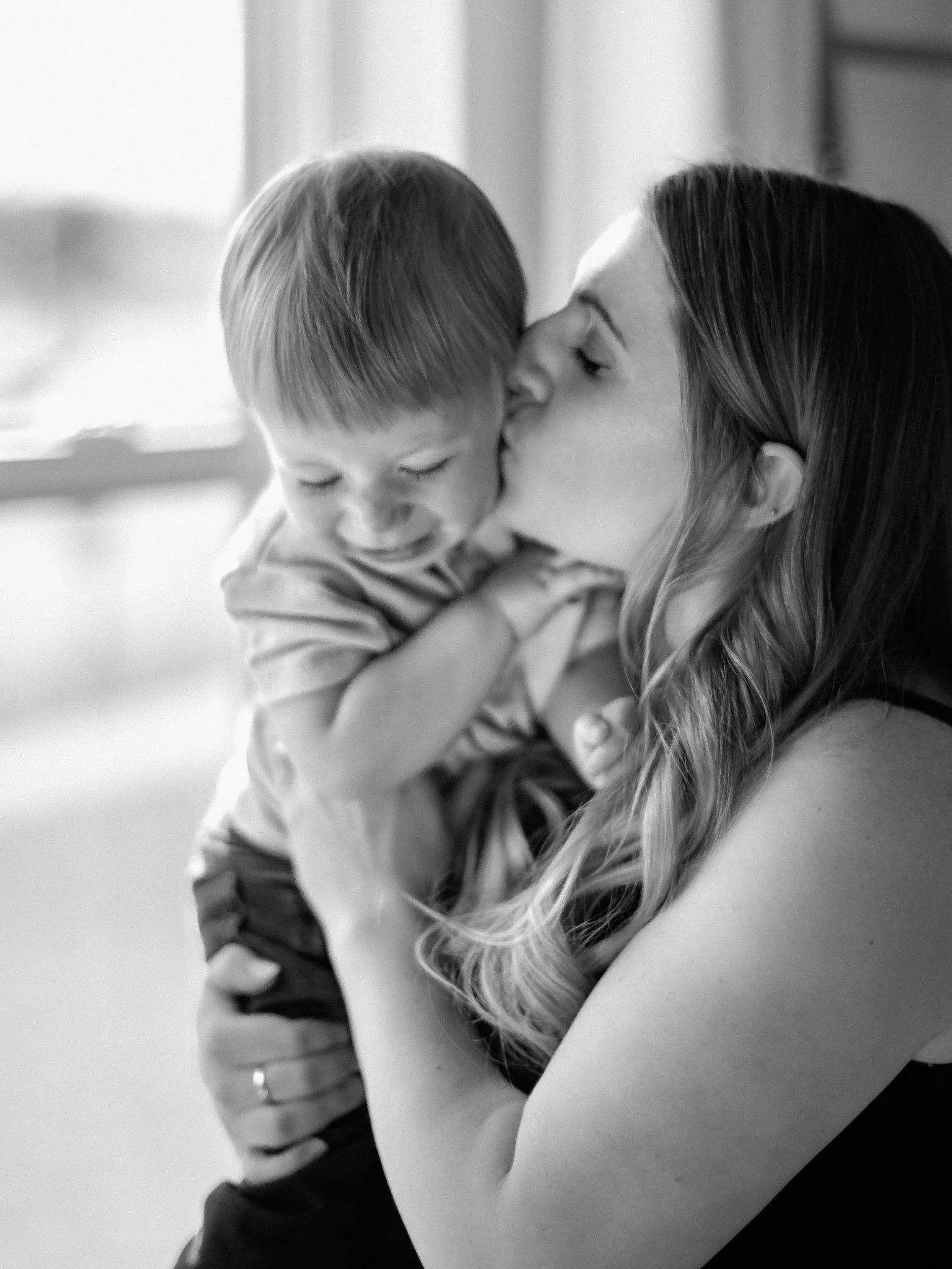 A woman gives a young boy a gentle kiss on the cheek, with the boy smiling and leaning into her affection, in a softly lit indoor setting.