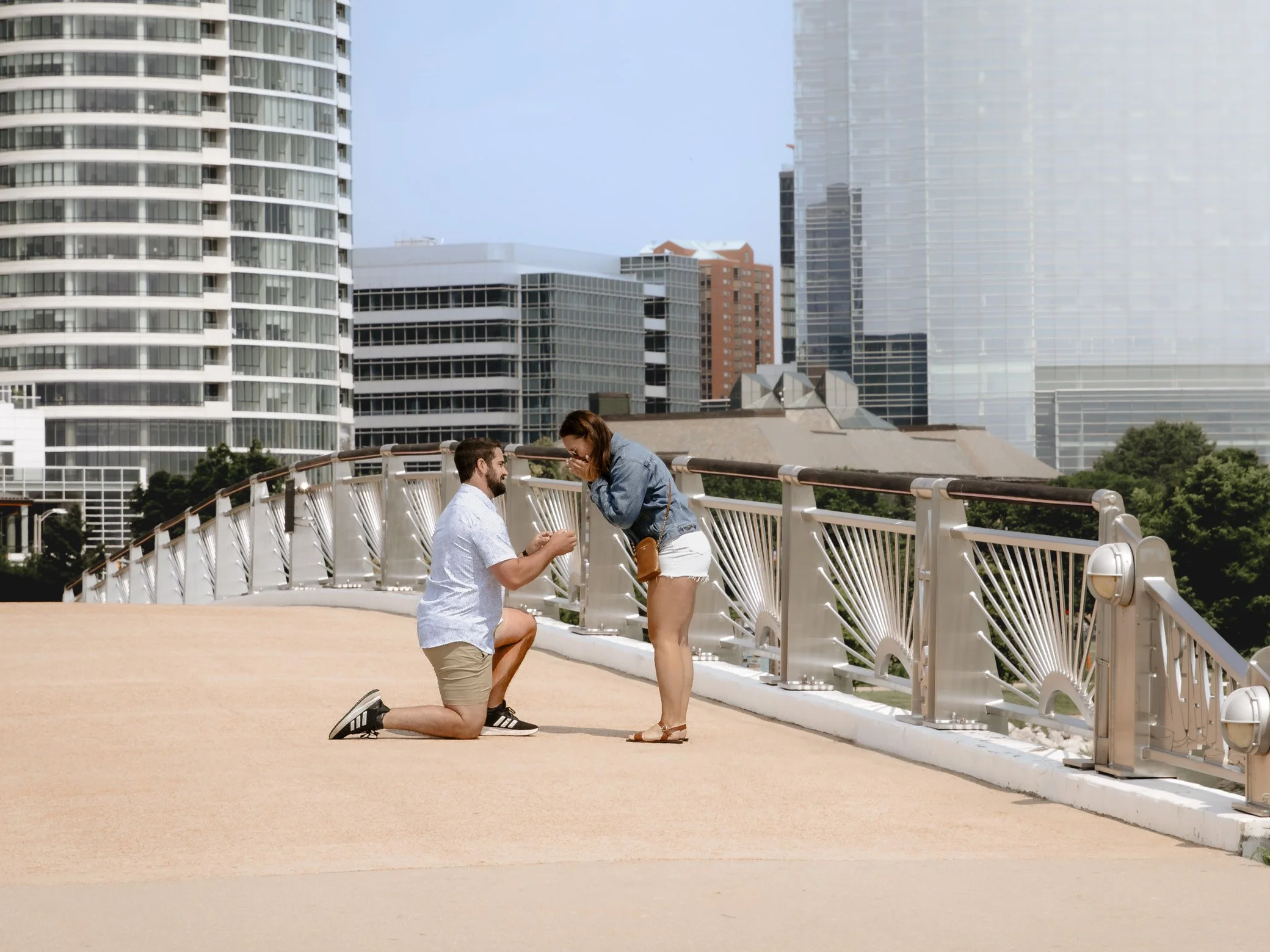A man proposing marriage to a woman on a city bridge with skyscrapers in the background.