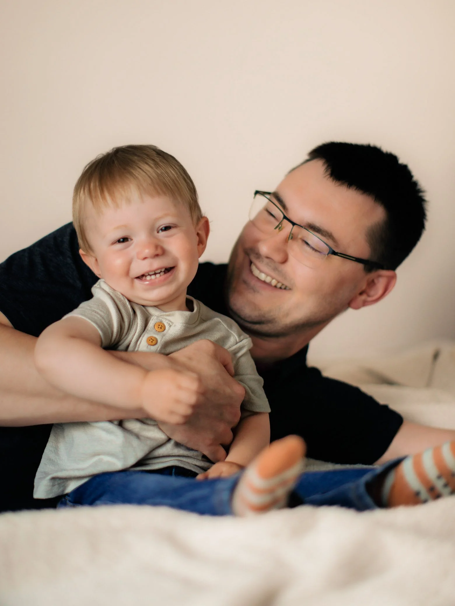 A smiling toddler with short light brown hair sitting on a bed, being held playfully by an adult man with black hair, glasses, and a black shirt, both appearing happy.