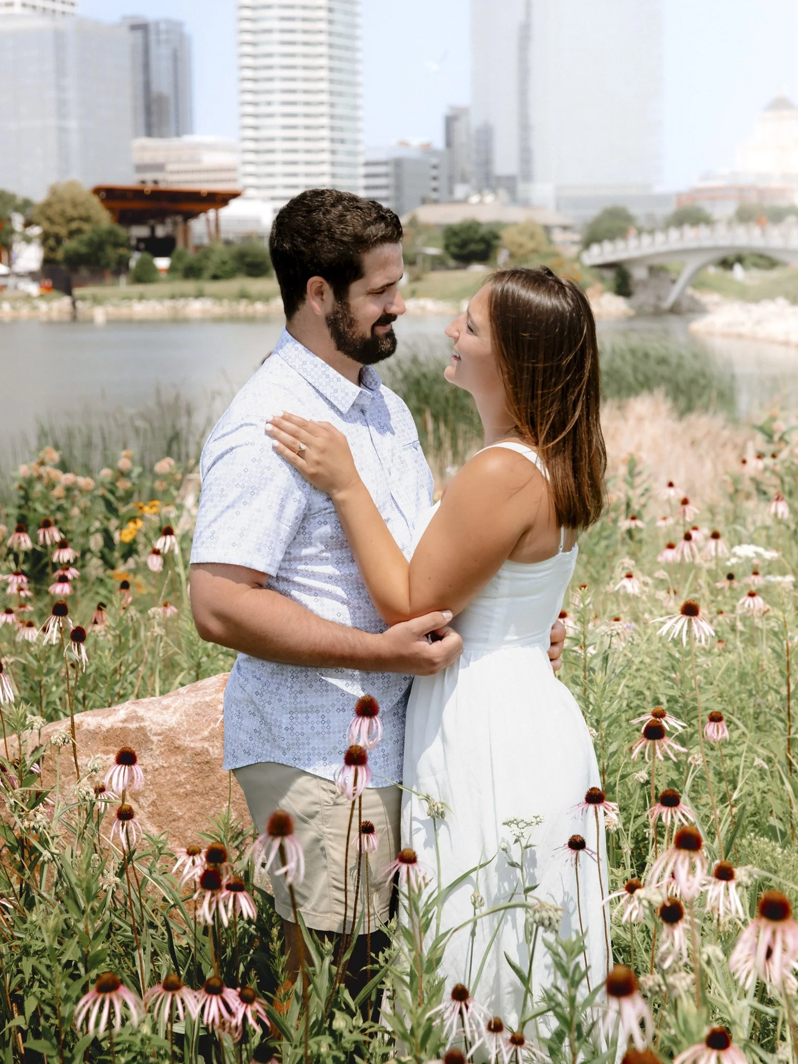 A couple embracing each other in a park filled with pink coneflowers, with a city skyline including tall buildings and a river in the background.