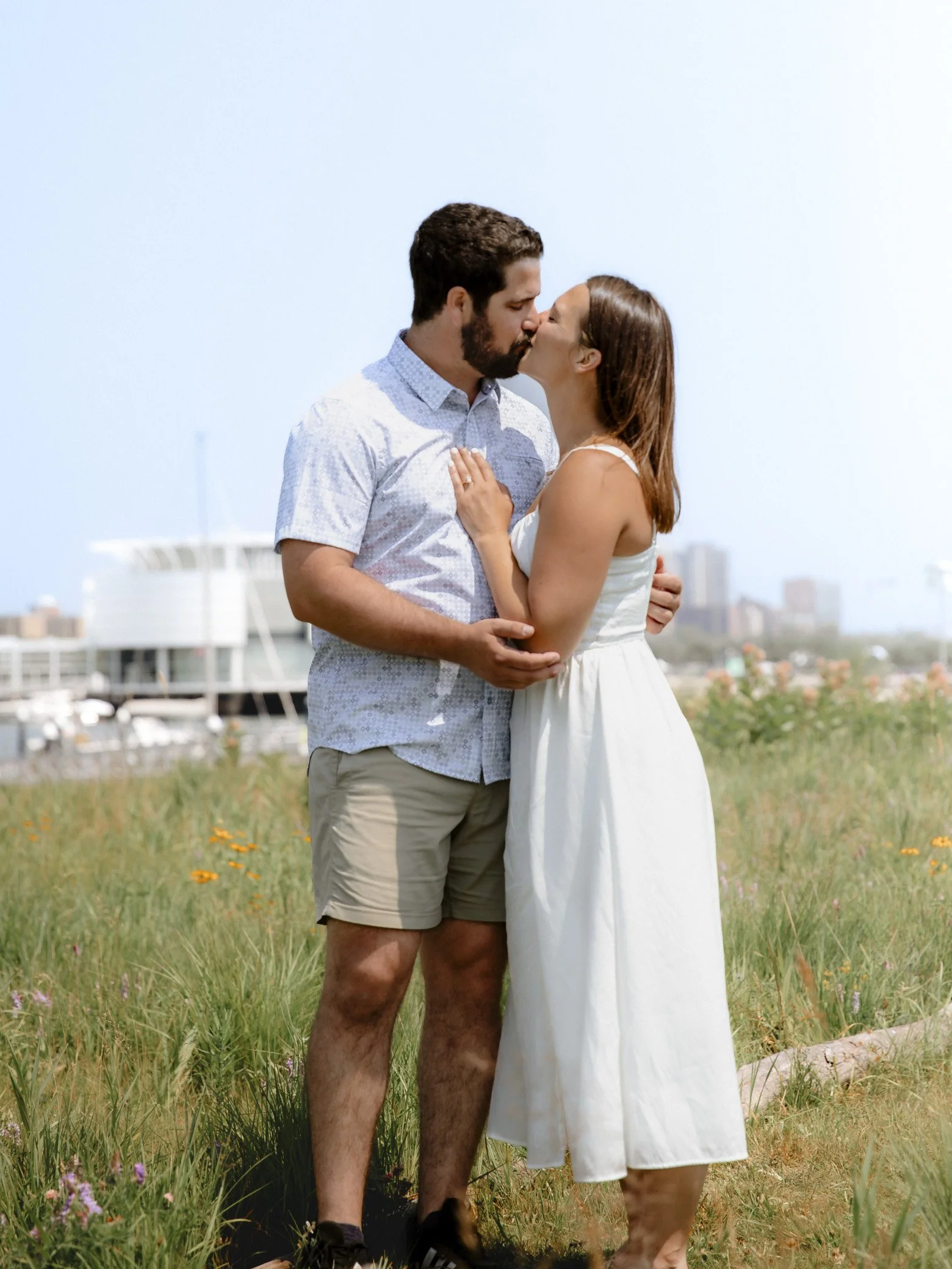 A couple sharing a kiss outdoors on a grassy field with a cityscape in the background.