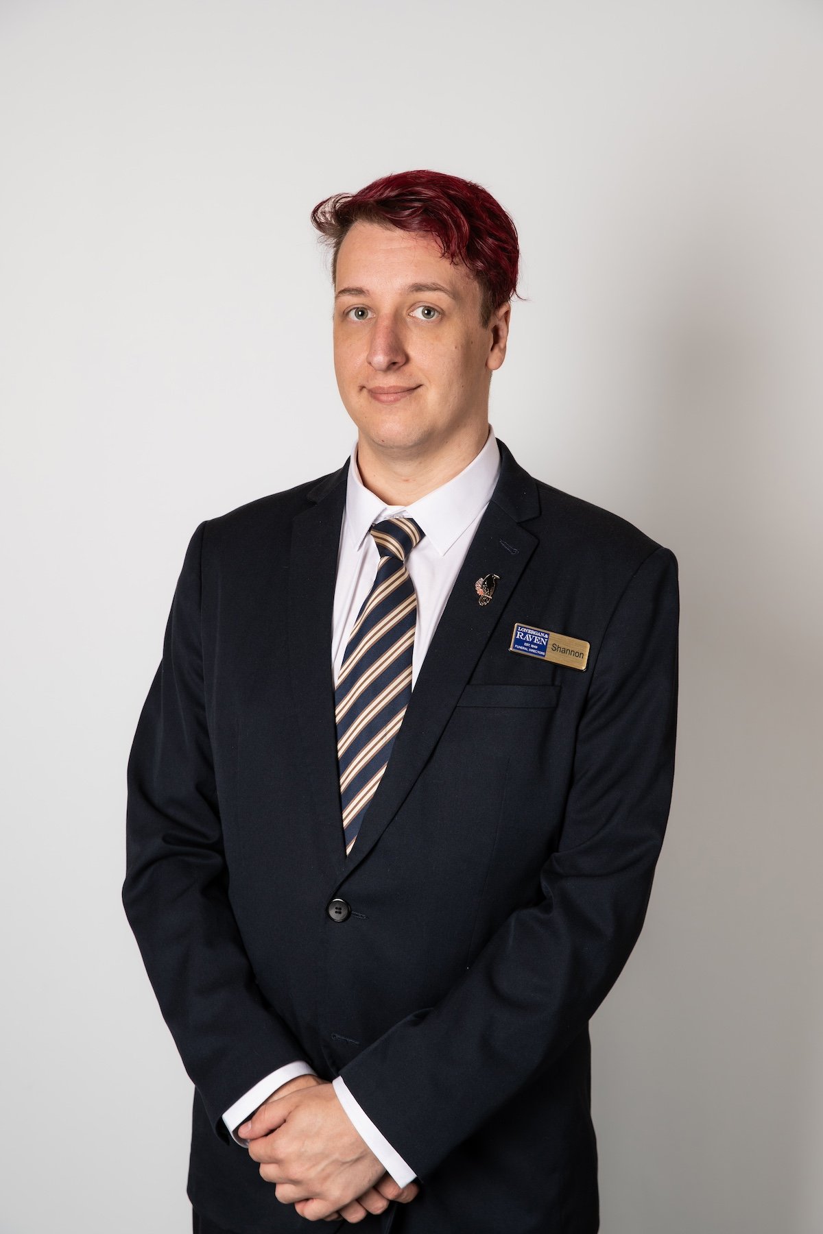 A young man with reddish hair in a formal suit and striped tie, standing with hands clasped in front, wearing a name tag that says 'Shannon,' against a plain white background.