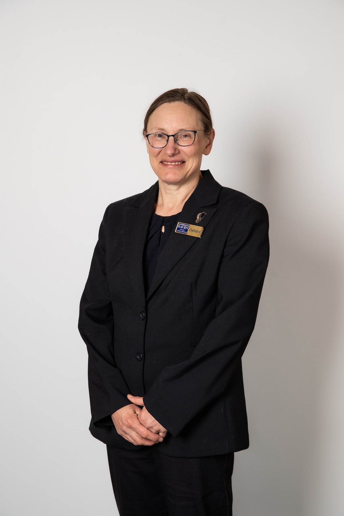 A woman in a black blazer and glasses, smiling, with a name tag on her lapel, standing against a plain white background.